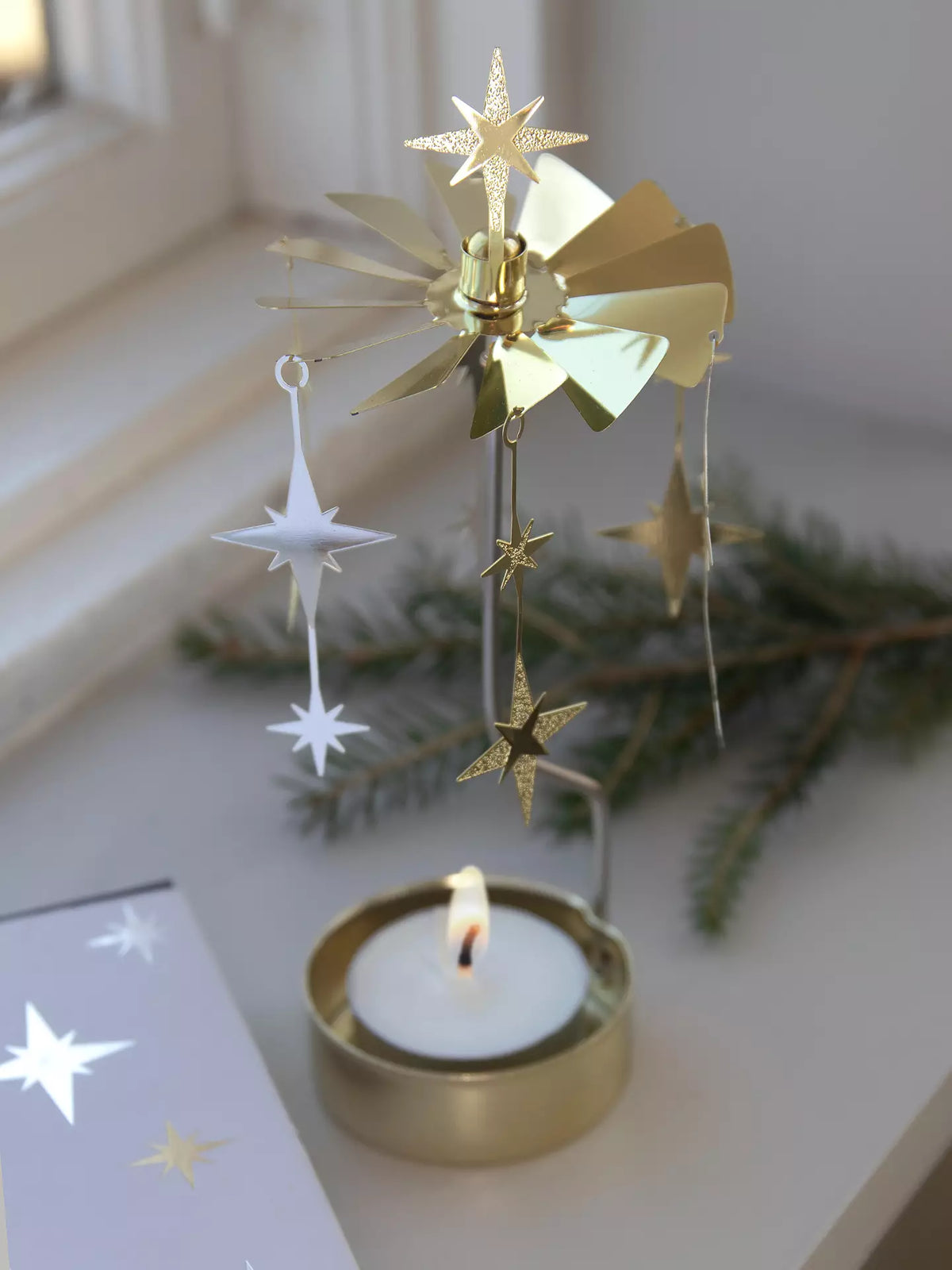 The Tealight Spinner, Sparkle in Gold and Silver by Pluto Design Sweden features spinning star ornaments and serves as a charming tealight carousel. Positioned by a window with a lit tealight candle at its base and a pine branch in the background, it makes an ideal piece of home decor or a thoughtful gift.