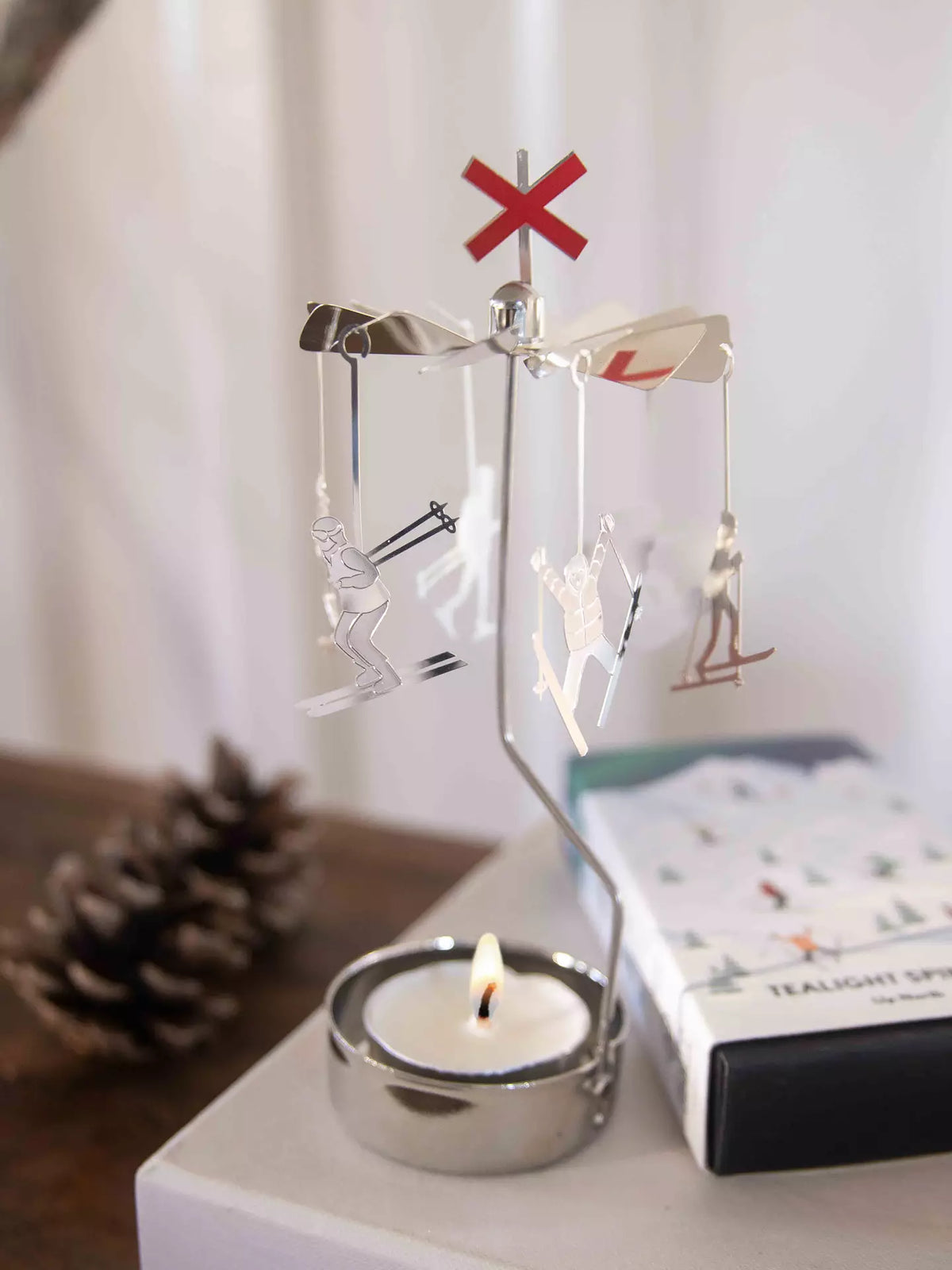 The Tealight Spinner, Up North in Silver by Pluto Design Sweden features dangling skier ornaments that rotate above a glowing tealight, topped with a distinctive red X sign. Nearby, a pinecone and box complement this charming and peaceful tealight spinner display.