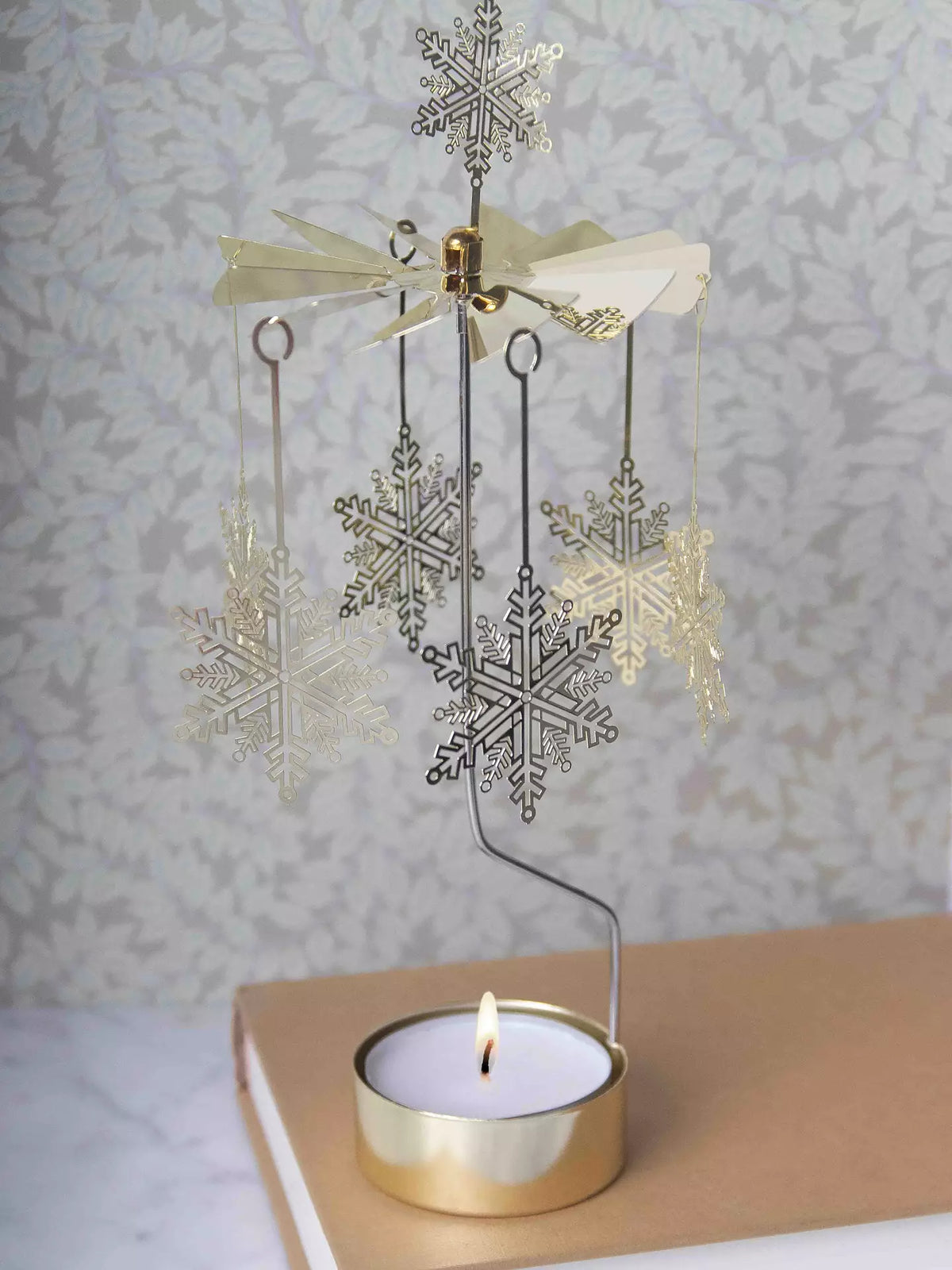 The Tealight Spinner, Big Snowstar in Gold by Pluto Design Sweden elegantly spins above a glowing tealight on a table, casting a magical light that enhances the festive Christmas mood. Perched on a closed book with a floral-patterned backdrop, this XL tealight holder instantly transforms any space into an enchanting winter wonderland.