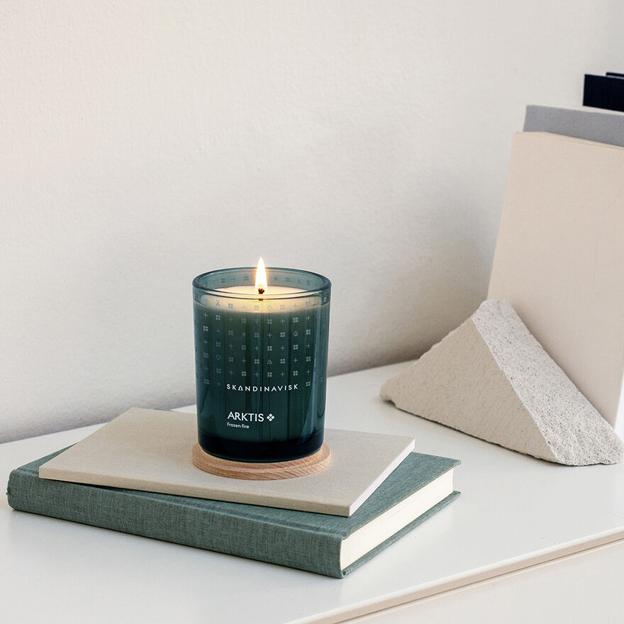 A lit Skandinavisk Scented Candle Arktis 50h, 200g in a green glass jar rests on a stack of books on a white surface, beside a triangular stone and upright folders.