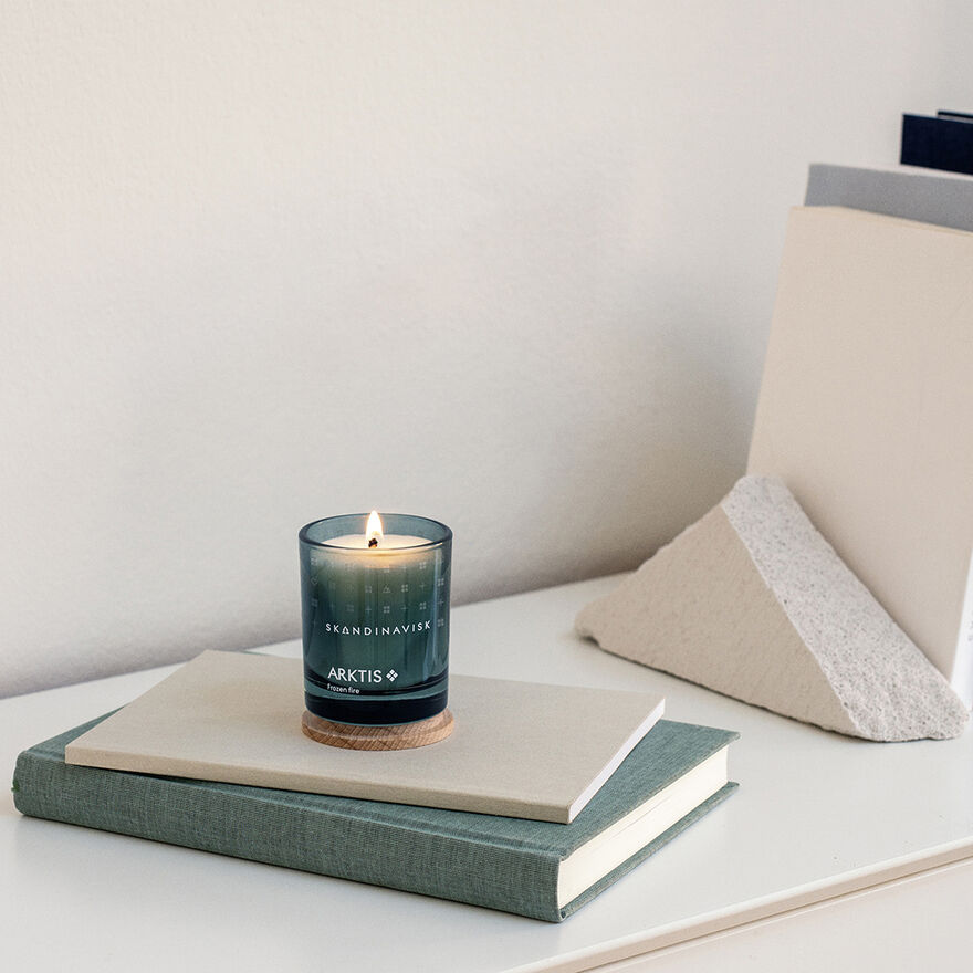 A Skandinavisk Mini Scented Candle Arktis, 65 g, glows atop a stack of books beside a triangular stone and upright folders on a white desk against a plain wall.