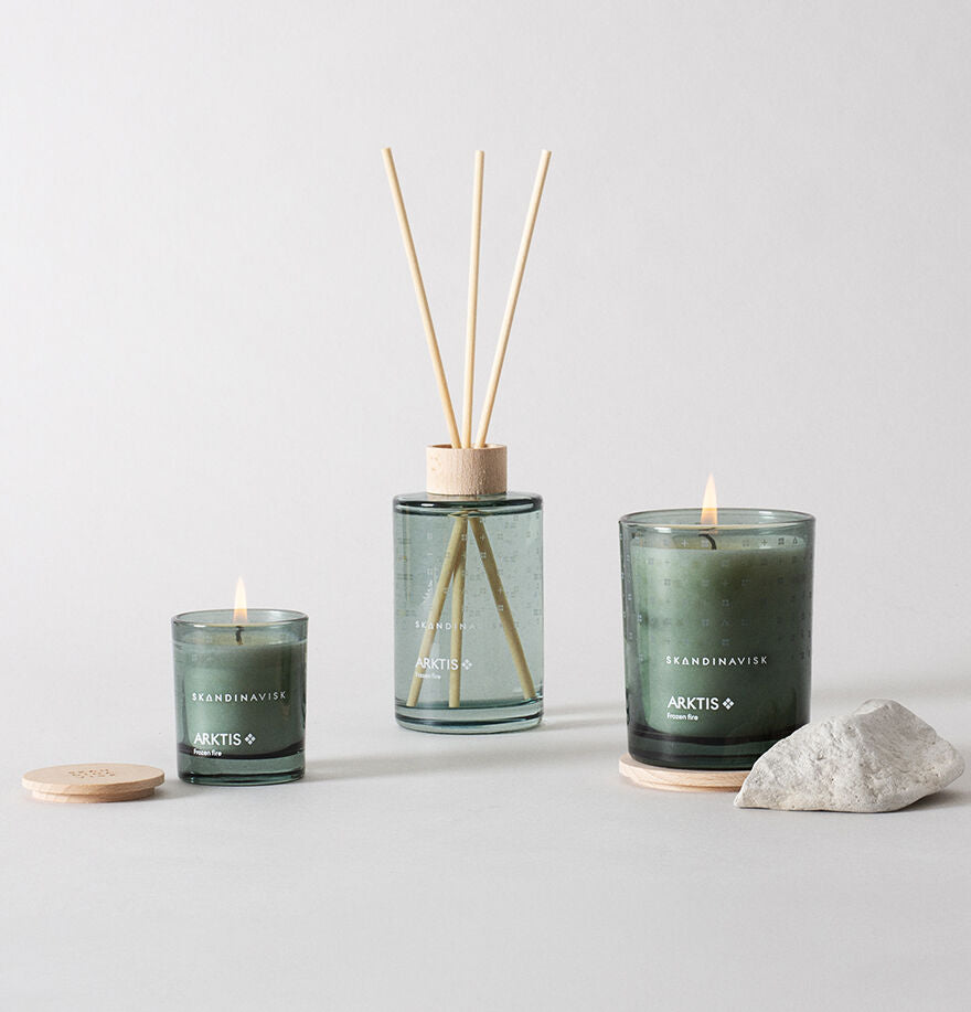 Three green-painted glass Mini Scented Candle Arktis, 65 g by Skandinavisk are displayed in holders alongside a reed diffuser with a wooden cap, all arranged on a light surface with nearby rocks and wooden lids. The SKANDINAVISK ARKTIS products feature arctic-inspired scented candles crafted from Swedish rapeseed wax.