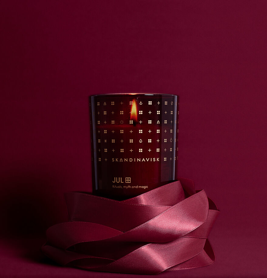 The Skandinavisk Seasonal Jul Scented Candle 50h, 200g, with its gingerbread aroma and deep red design, is adorned with a matching satin ribbon and set against a rich red backdrop for an authentic Scandinavian Christmas feel.
