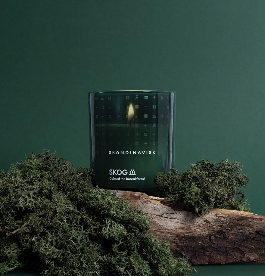 A lit Skandinavisk Scented Candle Skog 50h, 200g, made from Swedish rapeseed wax, sits on mossy wood against a dark green background.