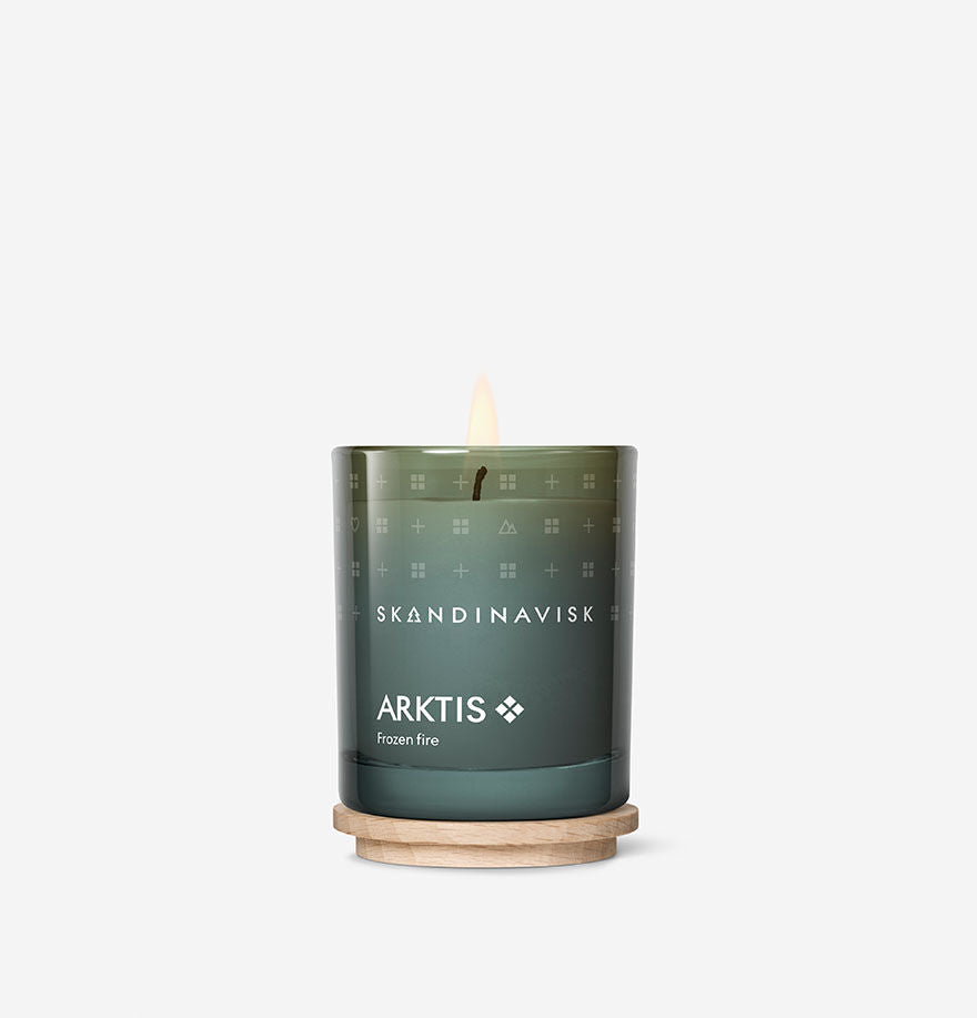 A lit Skandinavisk Mini Scented Candle Arktis, 65 g, crafted from Swedish rapeseed wax, glows in a green painted glass holder with white text and rests on a round wooden base against a plain white background.
