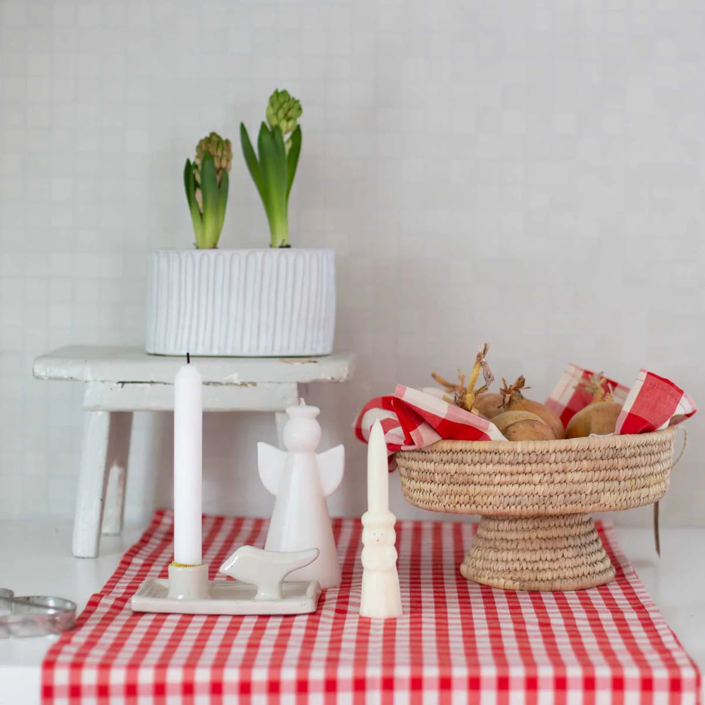 A handmade Candle Holder Earth Bird by A World of Craft with a white candle, an angel figurine, and a white planter with green shoots sit on a shelf beside a basket of potatoes and red-checked napkins on a red gingham tablecloth.