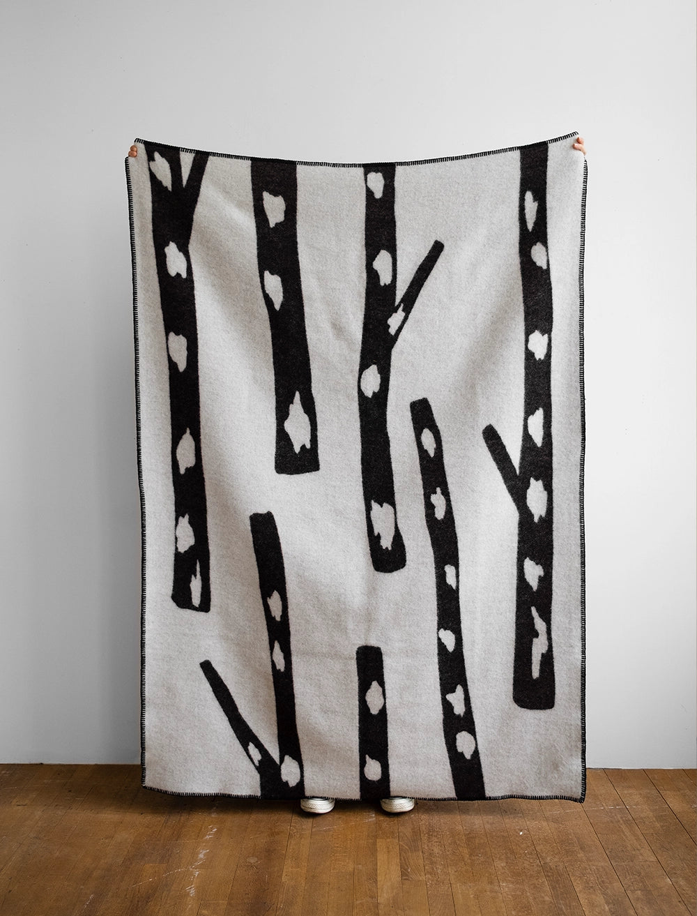 A person holds up the Fine Little Day Birch Wool Blanket, Reversible, in Black & White, featuring a Scandinavian-inspired birch pattern; only their shoes are visible below the cozy blanket.
