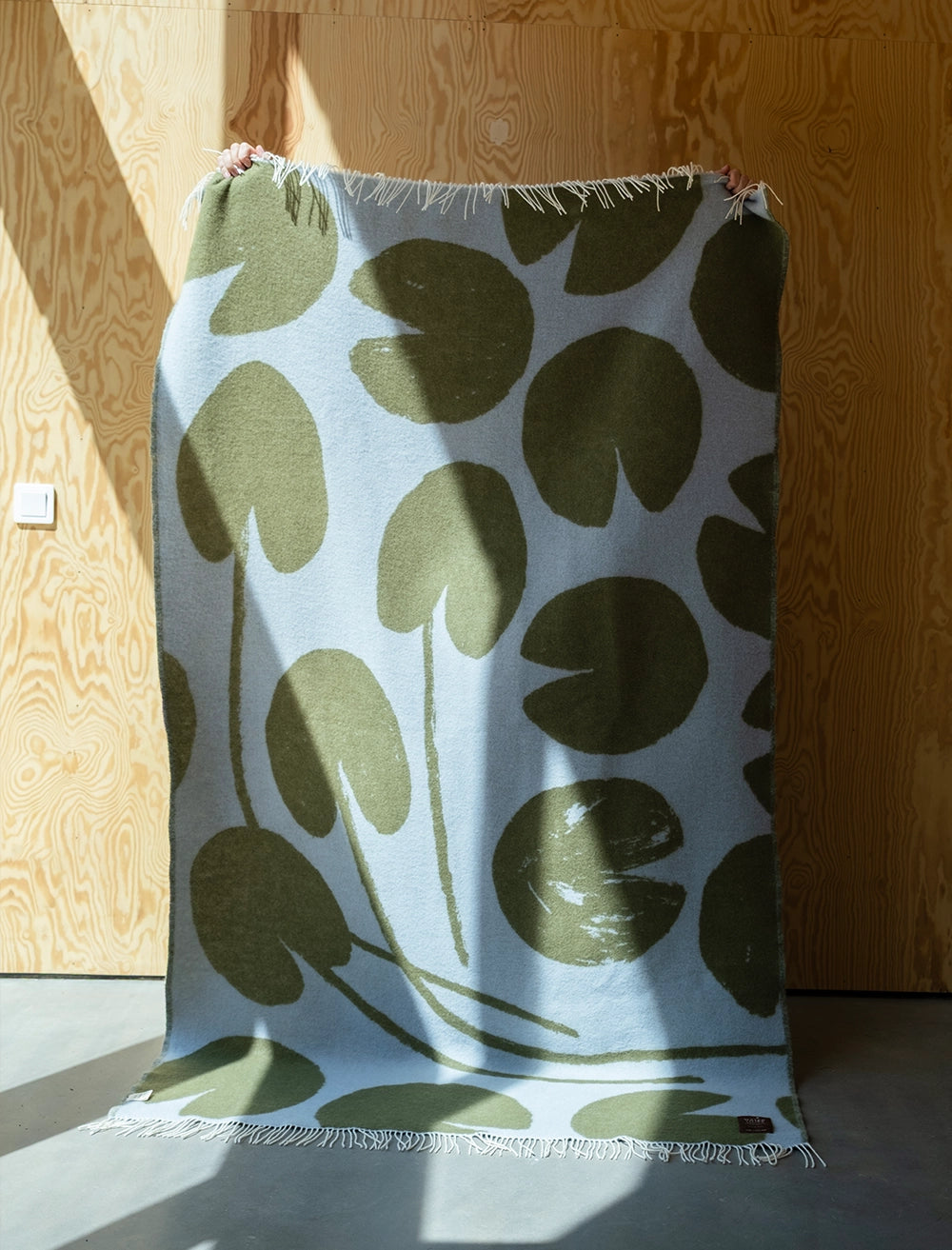 The Fine Little Day Water Lilies Wool Extra Large Blanket/Bedspread in reversible blue and green is displayed against a wooden wall, with sunlight partially illuminating its vibrant pattern.
