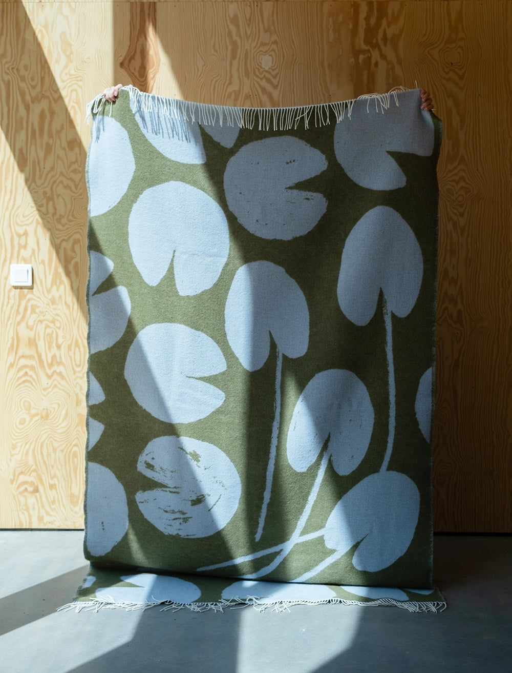 A person holds up the Fine Little Day "Water Lilies" Wool Extra Large Reversible Blanket/Bedspread in blue & green, showing its abstract leaf pattern, inspired by Water Lilies, against a wooden wall.