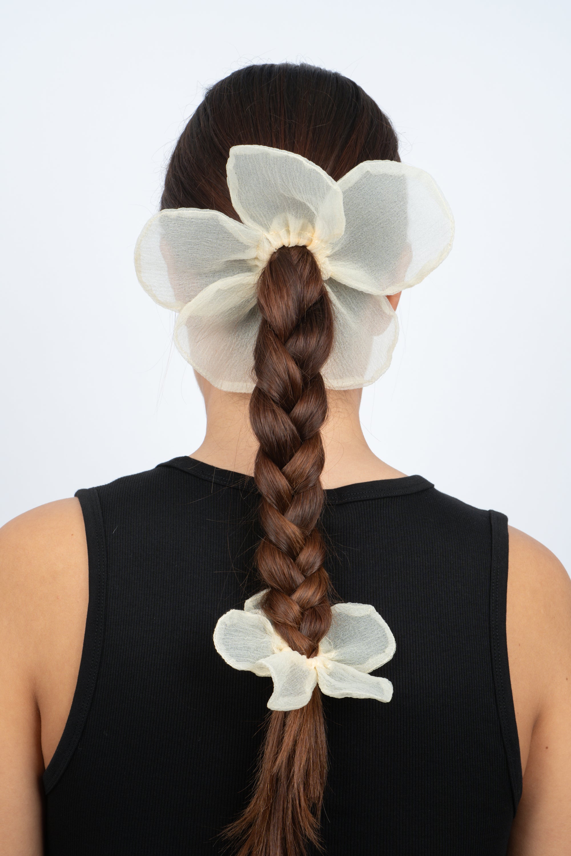 A woman with long brown hair in a braid wears a sleeveless black top. Her braid is styled with two large, sheer yellow Sui Ava Buttercup Elastic Big scrunchies—one at the top and one at the bottom.
