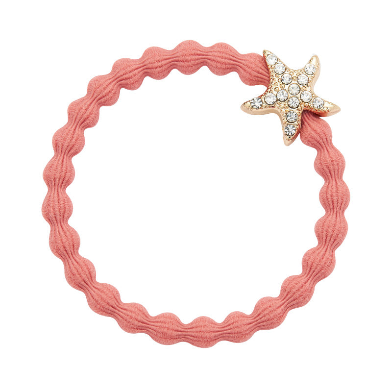 The ByEloise Bangle Band Starfish in Coral is a pink, wavy elastic hairband featuring a gold starfish charm embellished with sparkly rhinestones.