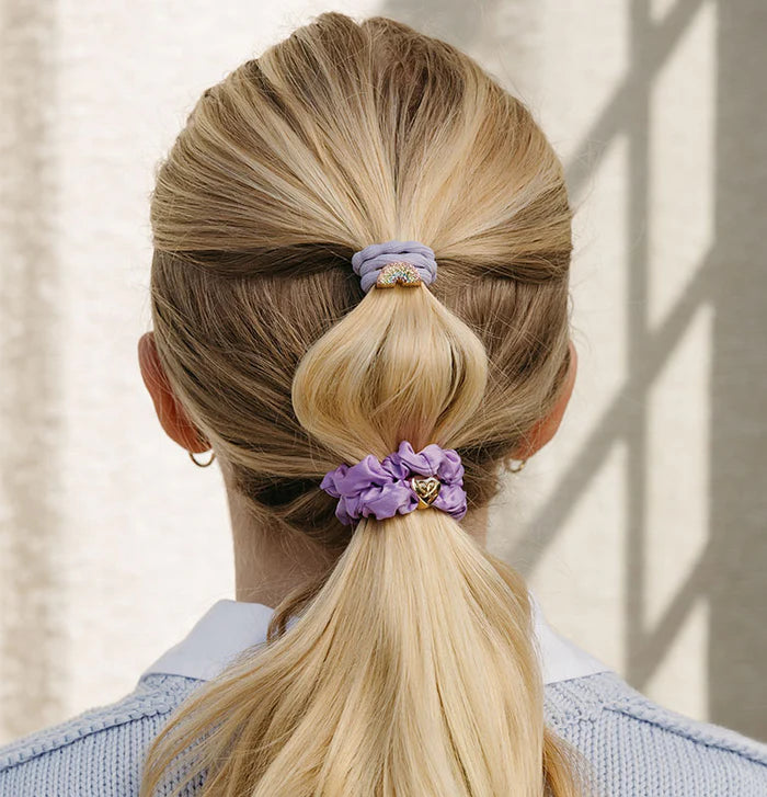 A woman with blonde hair in a ponytail, secured by the ByEloise Bangle Scrunchie set of 2 in lilac and purple (Lavender Skies), wears a light blue sweater and hoop earrings, facing away from the camera.