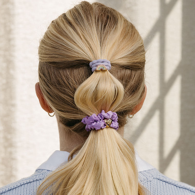 A woman with blonde hair in two ponytails, each tied with ByEloise Bangle Band Bling Rainbow and Lavender hair ties—one has a jewel accent. She is dressed in a light blue sweater over a collared shirt.
