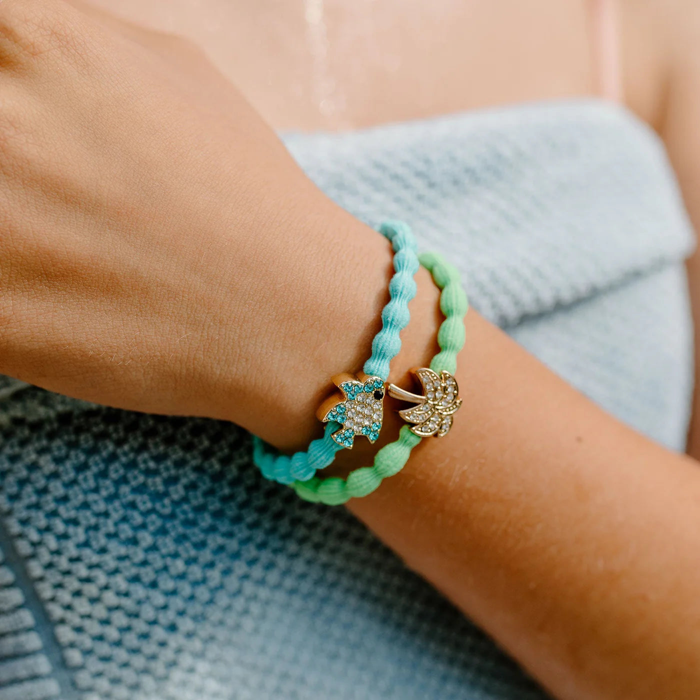 A person wearing a light blue textured top has two braided bracelets, one blue and one green, each featuring jeweled animal charms, on their wrist. Their hair is pulled back with the ByEloise Bangle Band in Neon Green, adorned with a gold palm tree charm, adding a playful touch to the look.