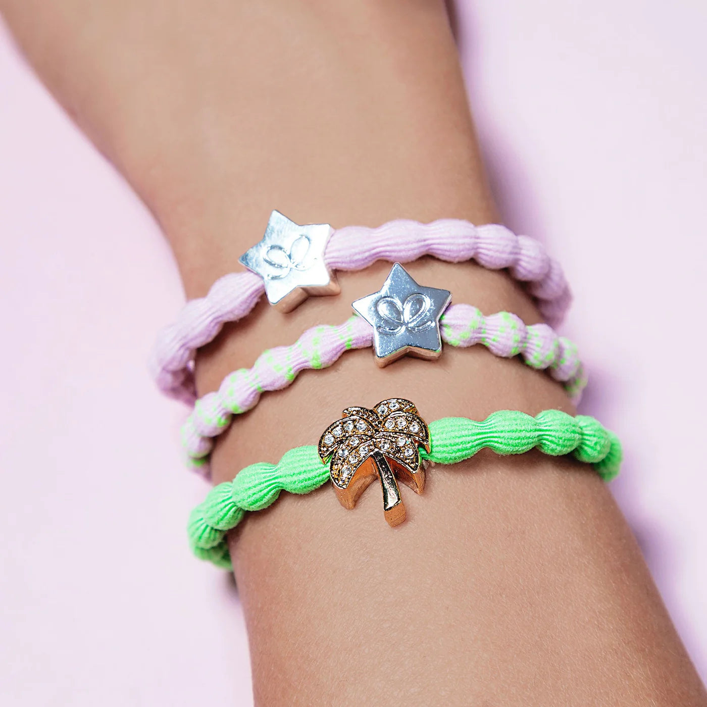 A wrist adorned with three braided bracelets—a pair of lavender bands each featuring a silver star charm, and the ByEloise Bangle Band in Neon Green, Palm Tree, boasting a gold palm tree charm accented with small rhinestones reminiscent of a gold palm tree hairband—set against a light pink background.