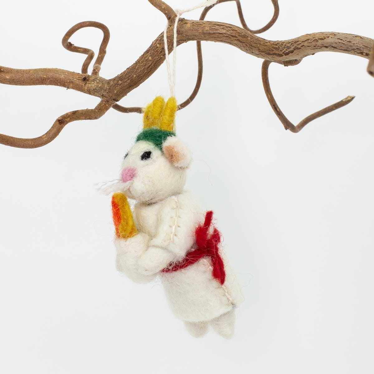 Handmade in wool by A World of Craft, the Lucia Christmas Ornament features a felt mouse dressed in a white outfit with a red sash, honoring Swedish Santa Lucia tradition. This charming ornament, holding a carrot, hangs delicately from a brown twisted branch against a crisp white background.