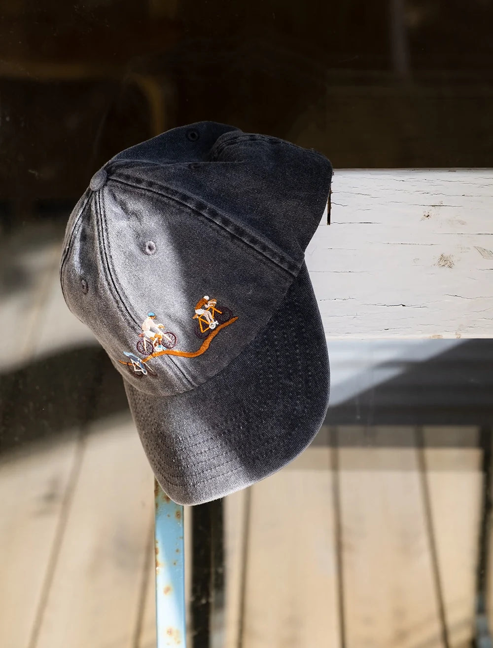 The Giro Bike Cap Embroidered by Fine Little Day, in vintage black, featuring embroidered cyclists in orange, white, and blue, hangs on a weathered white wooden railing bathed in sunlight.