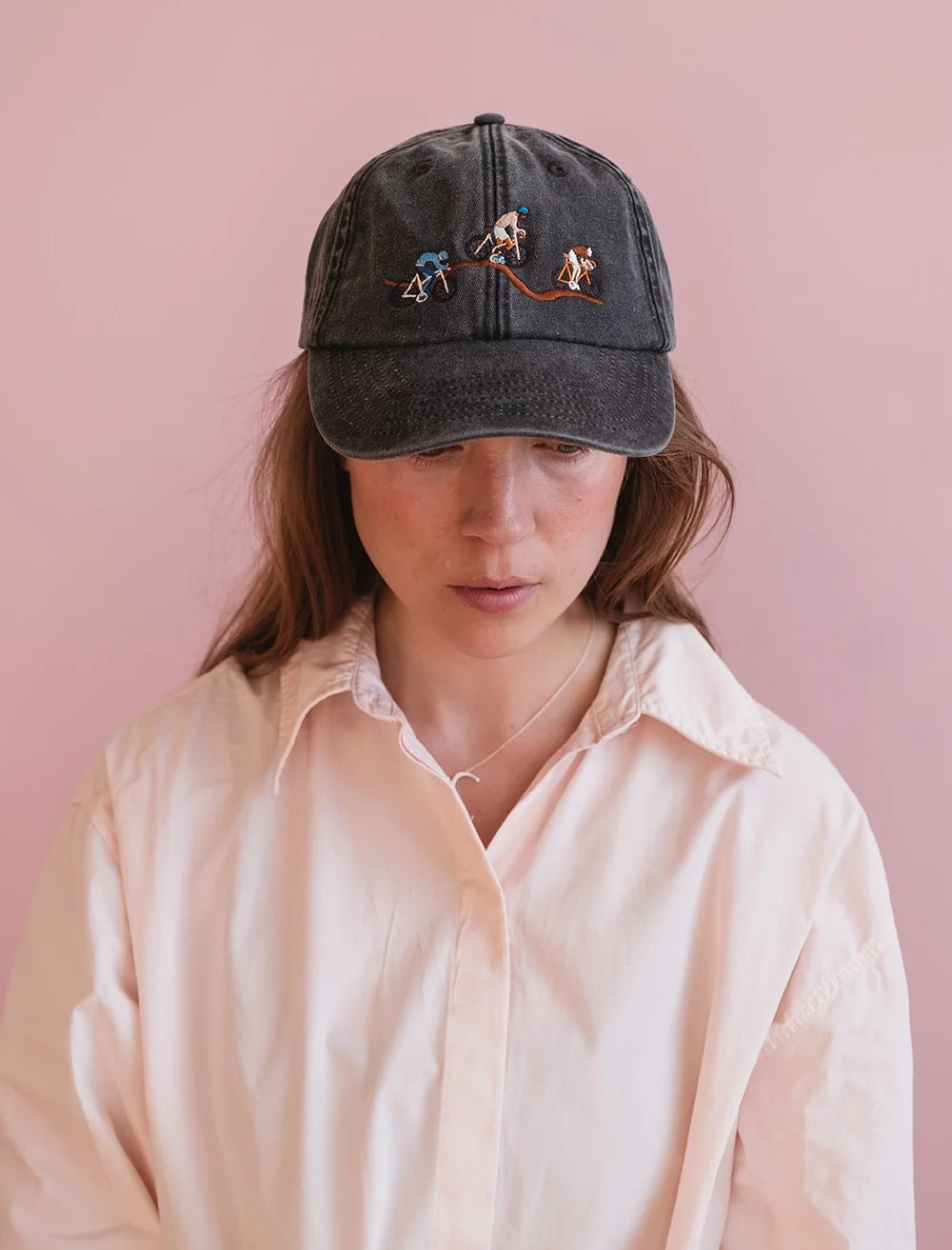 A person in a pale pink shirt wears the Fine Little Day Giro Bike Cap Embroidered in Vintage Black, featuring small, colorful embroidered figures on the front, standing against a light pink background.