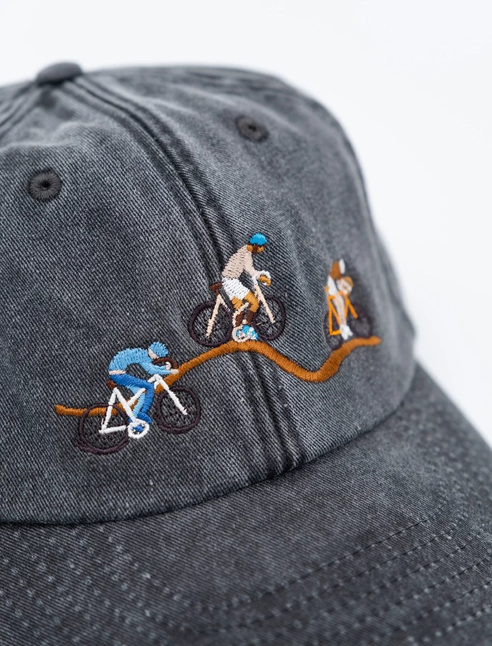 A close-up of the Fine Little Day Giro Bike Cap Embroidered in Vintage Black—a 6-panel strapback hat adorned on the front with embroidered cyclists riding along a winding brown path, capturing the spirit of the Giro d’Italia.