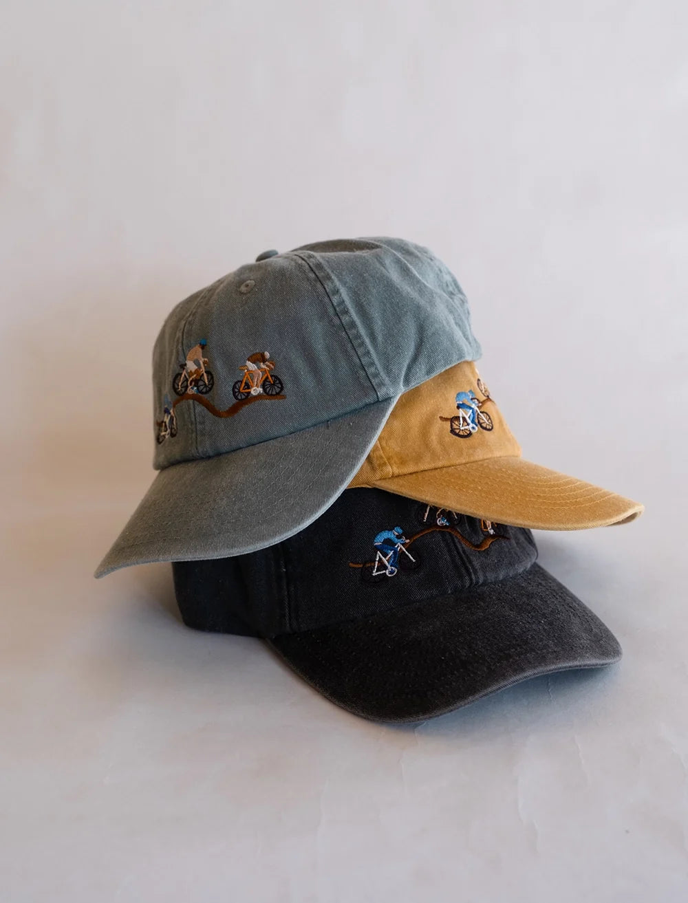 Three Fine Little Day Giro Bike Caps in Vintage Black, Gray, and Yellow are stacked atop one another. Each cap is a strapback 6-panel design, featuring small embroidered figures riding bicycles on the front—a nod to the Giro d’Italia—set against a plain light background.