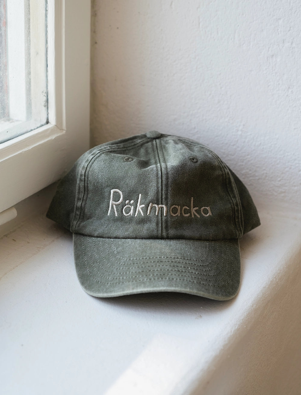 The Räkmacka Cap Embroidered in Cotton, Vintage Olive (Shrimp Sandwich) by Fine Little Day rests on a white windowsill.