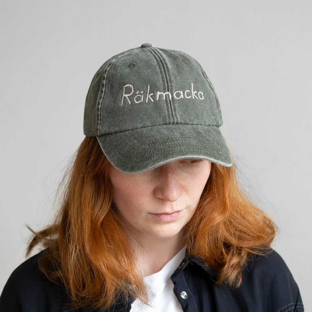 A person with long red hair wears the Fine Little Day Räkmacka Cap, embroidered in cotton (Shrimp Sandwich), vintage olive, paired with a black shirt over a white top against a plain background.