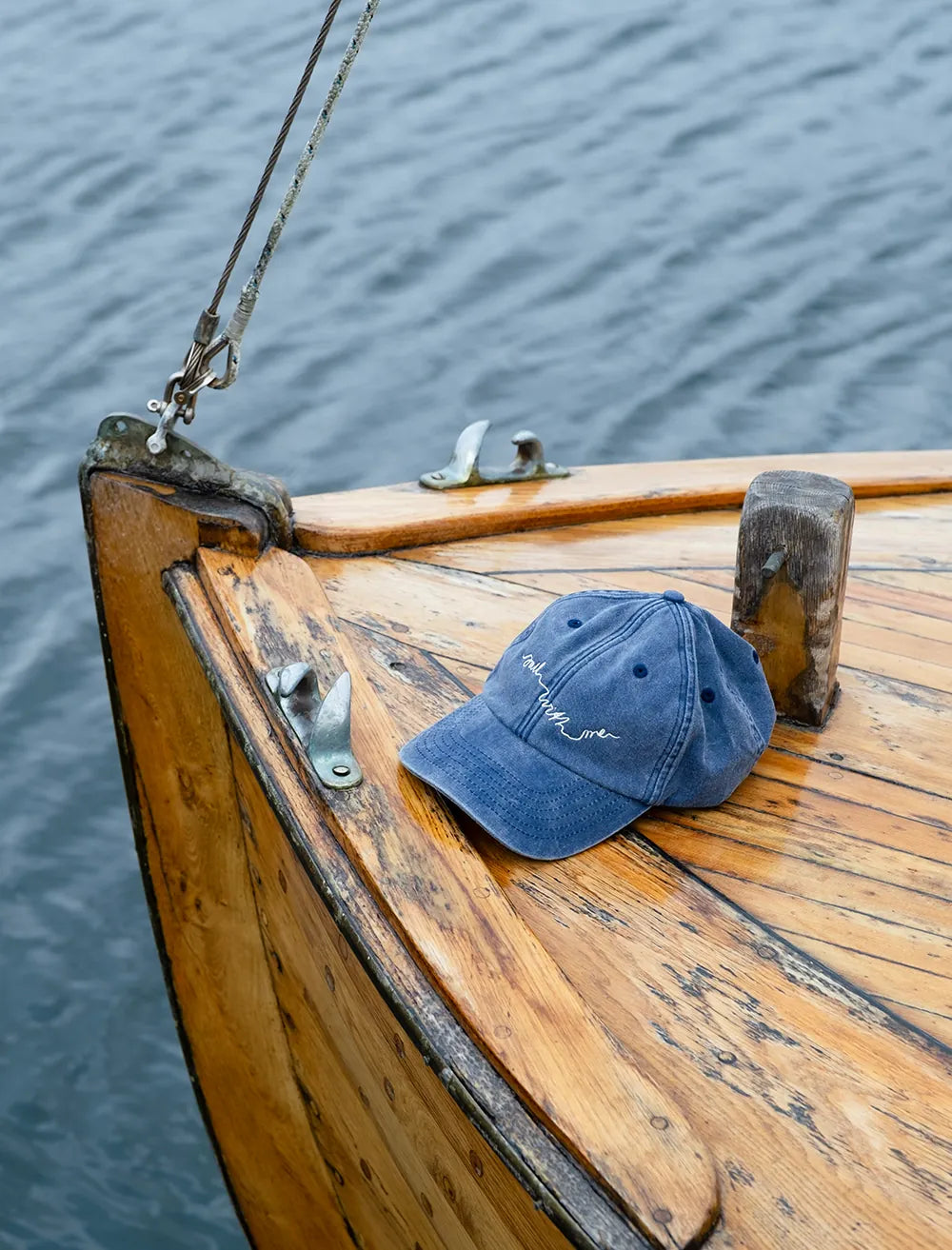 The Sail With Me Cap Embroidered in vintage denim by Fine Little Day sits on the boat’s wooden bow, inviting you to set sail across calm waters.