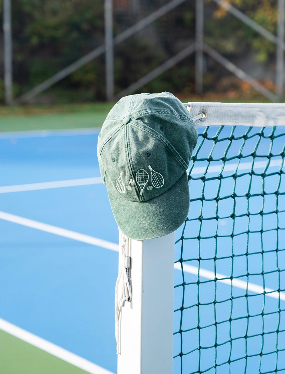 The Fine Little Day Tennis Cap Embroidered in Cotton, Vintage Green, featuring tennis rackets and an adjustable strap, hangs on the net post of an outdoor tennis court.