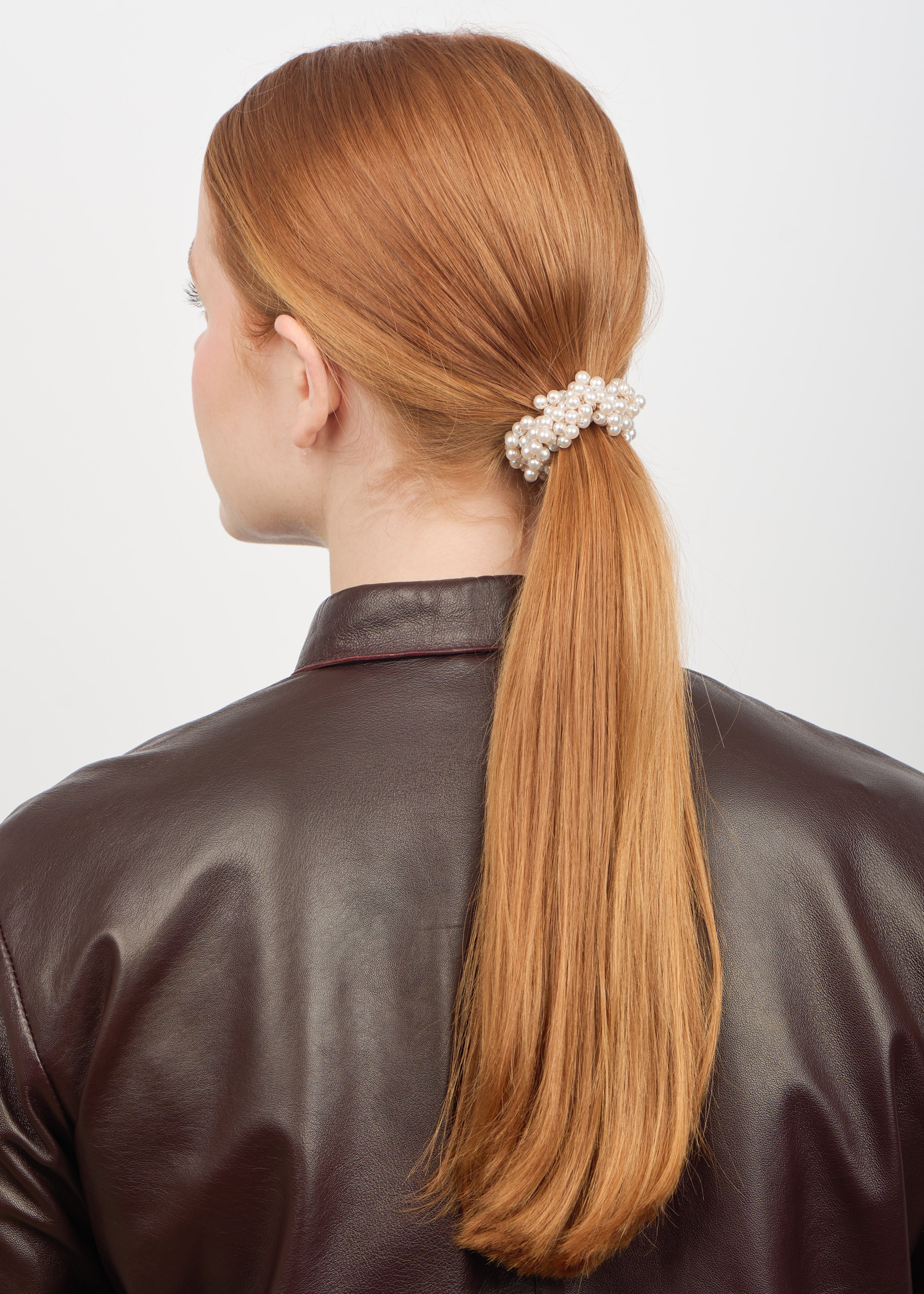 A woman with straight, long auburn hair in a low ponytail, secured with the Sui Ava Classic Pearl Elastic Hair Band in Beige, wears a dark brown leather jacket and is photographed from behind against a plain light background.
