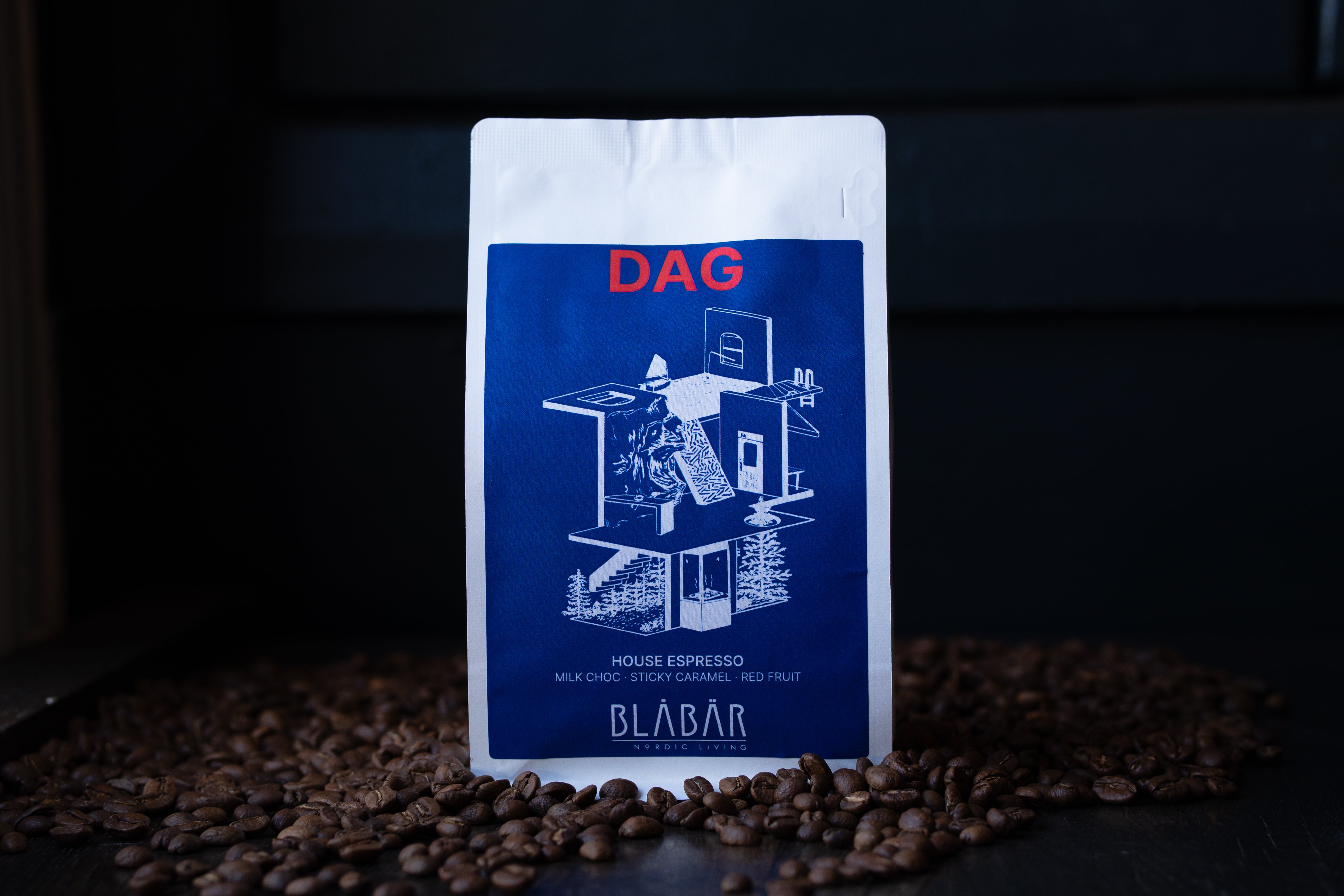 A white coffee bag labeled DAG from Blåbär Nordic Living stands upright on a dark surface, surrounded by scattered coffee beans. The packaging highlights the House Coffee Beans—Single Origin Colombia (250g)—with tasting notes of milk chocolate, sticky caramel, and red fruit, making it an inviting choice for those who appreciate a rich espresso or a smooth flat white.