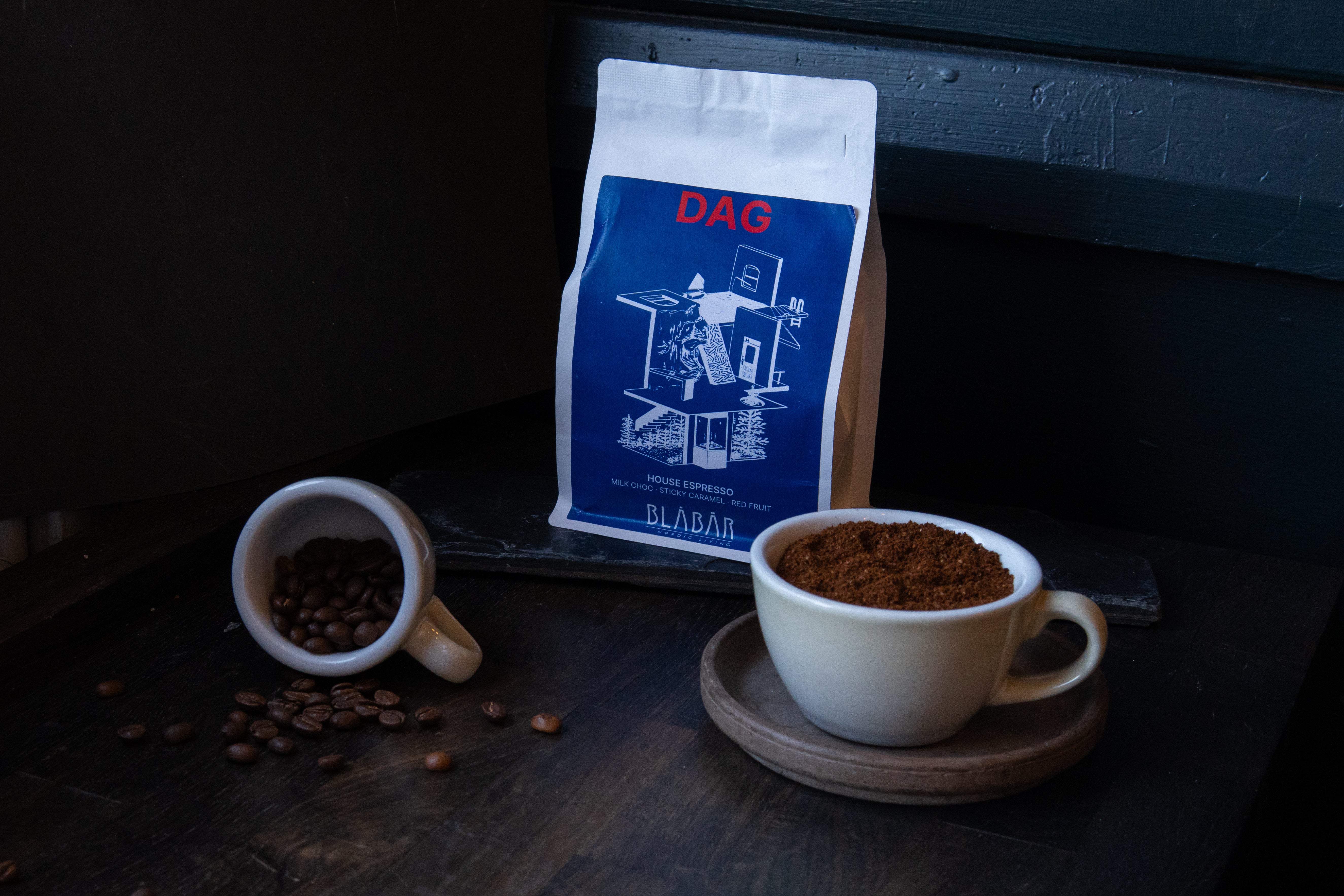 A bag of Blåbär Nordic Living DAG House Coffee Beans Blåbär, Single Origin Colombia, 250g stands behind a cup filled with ground espresso, with a tipped cup spilling coffee beans beside them, all set on a dark wooden surface.