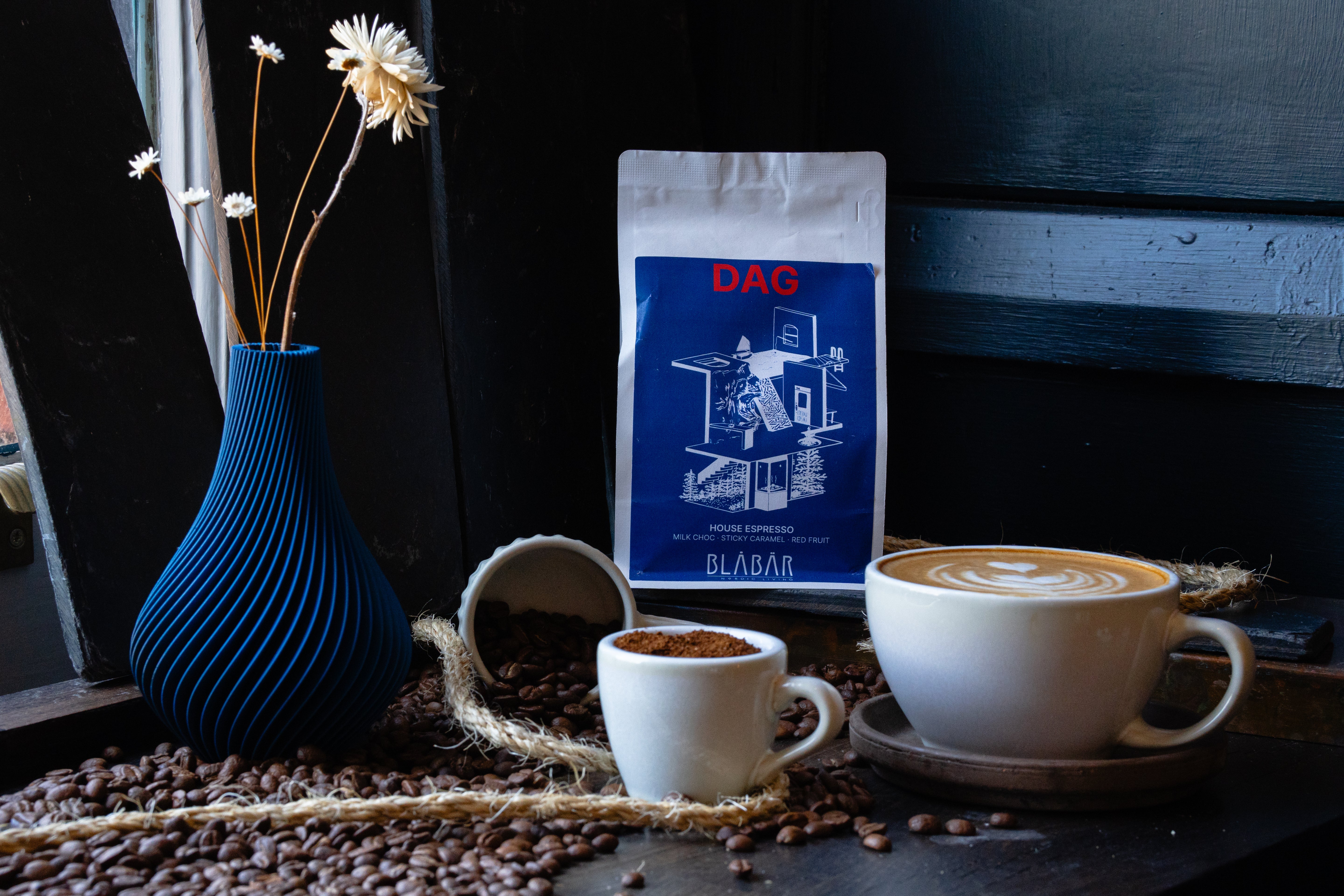 A latte topped with intricate latte art, a small cup of espresso, a blue vase filled with dried flowers, and a 250g bag of Blåbär Nordic Living’s DAG House Coffee Beans Blåbär (Single Origin Colombia) rest on a coffee bean-strewn table by the window—creating the perfect cozy scene for any coffee enthusiast.