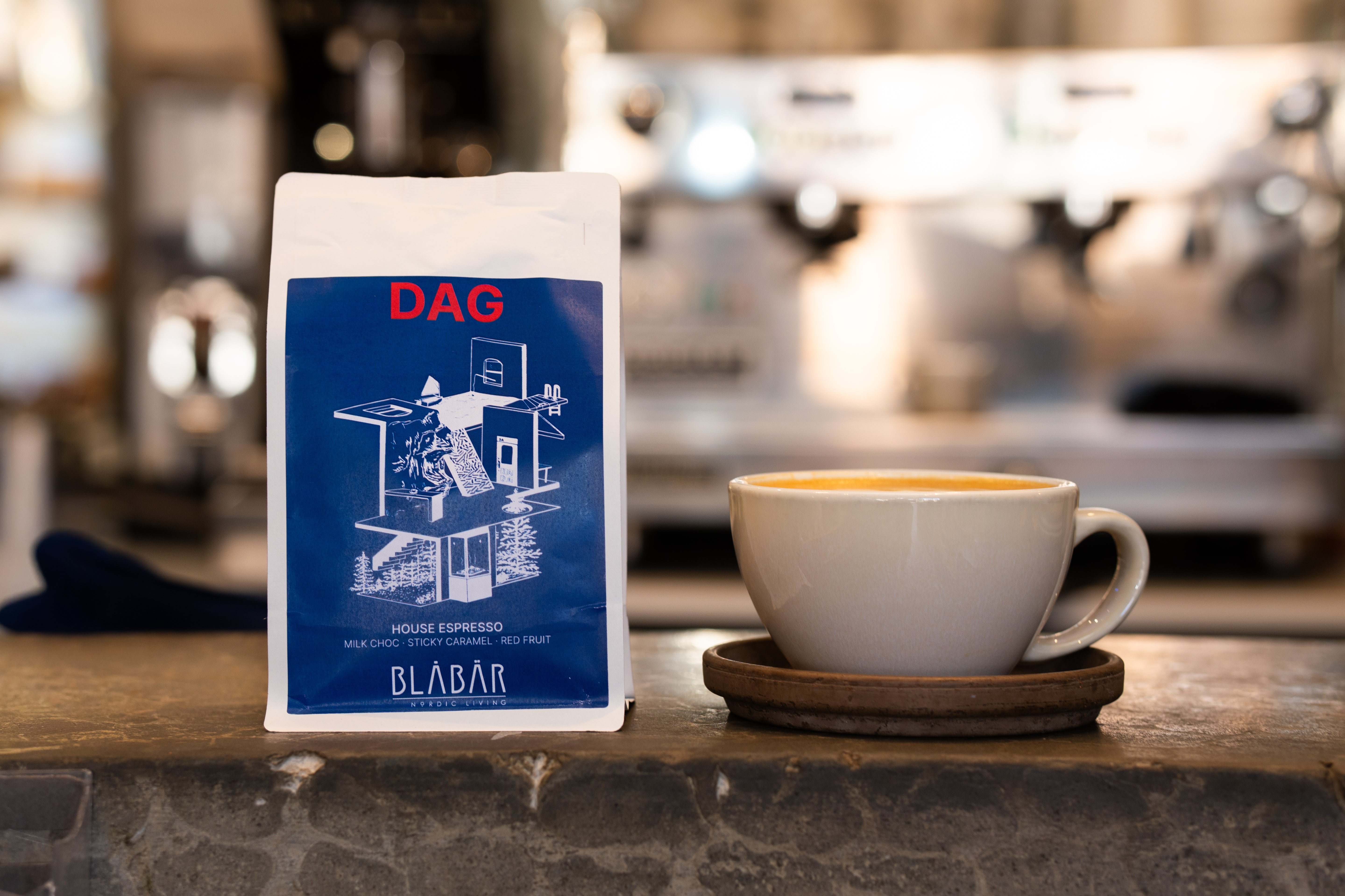 A white coffee cup on a saucer sits beside a blue and white bag of Blåbär Nordic Living DAG House Coffee Beans Blåbär, Single Origin Colombia, 250g, on a stone countertop, with a blurred espresso machine in the background—capturing the inviting moment before enjoying a rich flat white or your favorite espresso.
