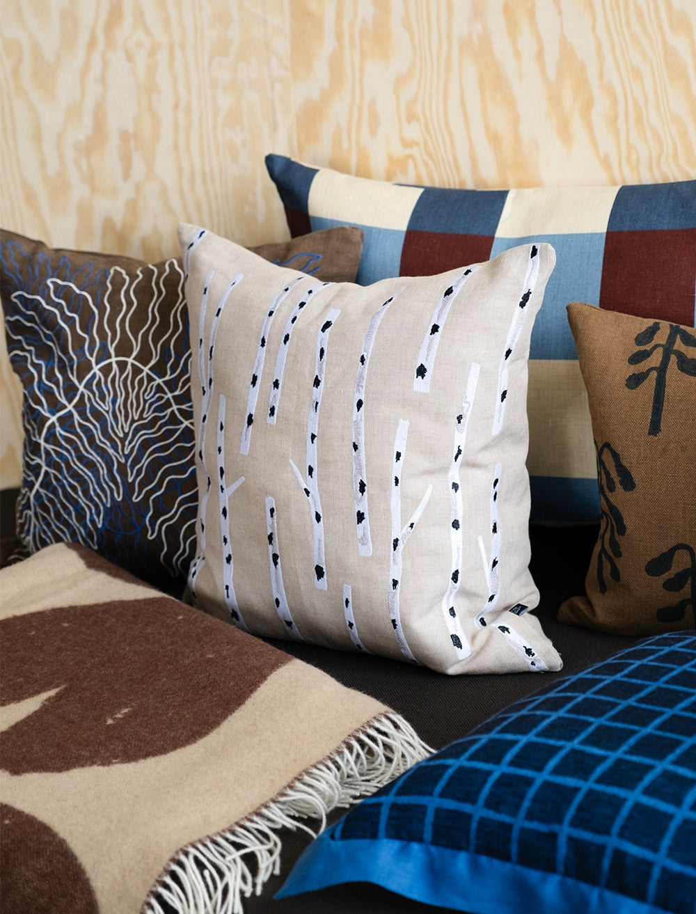 A group of patterned pillows, including the Fine Little Day Birch Embroidered Cushion Cover in Nature, and a blanket are arranged on a dark sofa in front of a light wood-paneled wall.