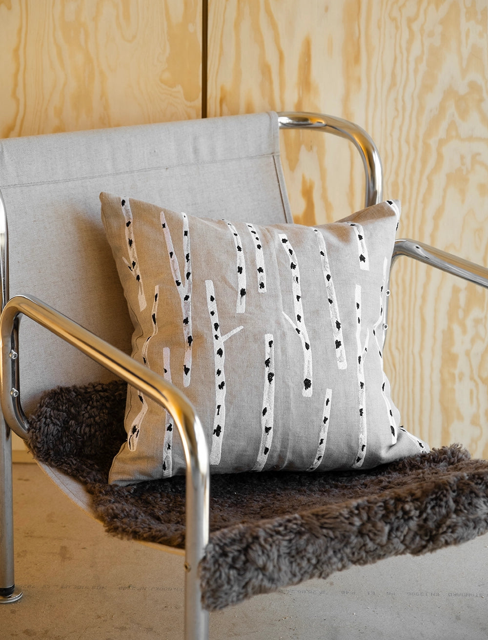 The Birch Embroidered Cushion Cover in Nature by Fine Little Day, featuring a neutral birch tree design, is placed on a modern metal chair with a gray seat and brown faux fur throw.