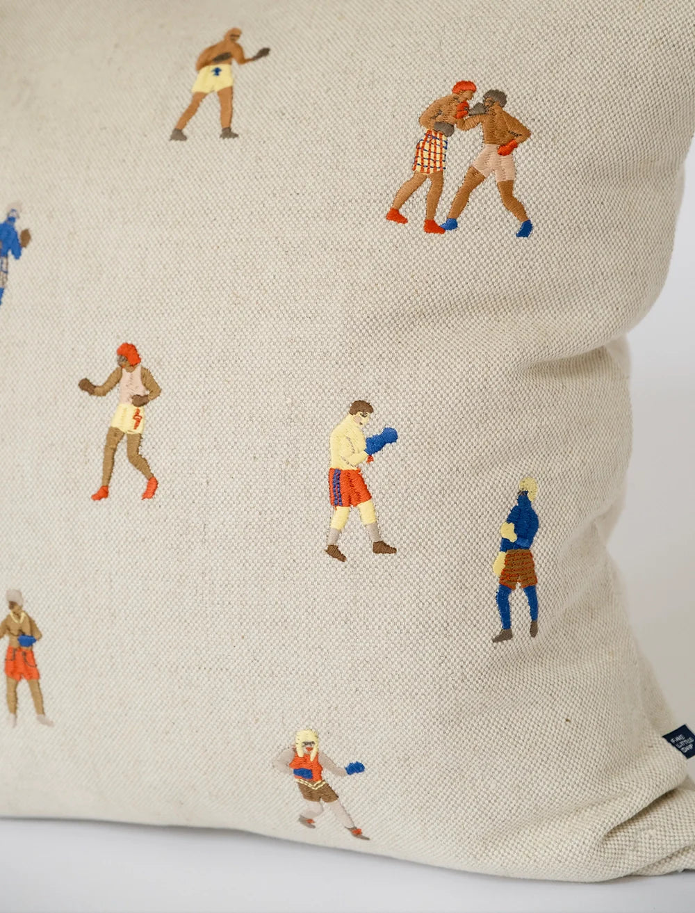 The Boxers Embroidered Cushion Cover by Fine Little Day is a beige linen cover featuring small, colorful embroidered boxers in different poses, each with unique trunks and gloves.