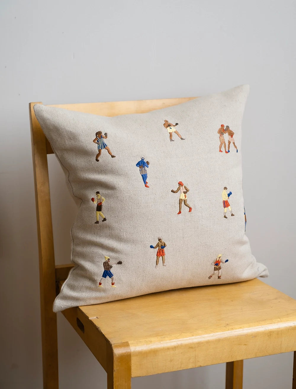 The Boxers Embroidered Cushion Cover by Fine Little Day, in beige linen with colorful boxer motifs, rests on a light wooden chair against a plain light gray wall.
