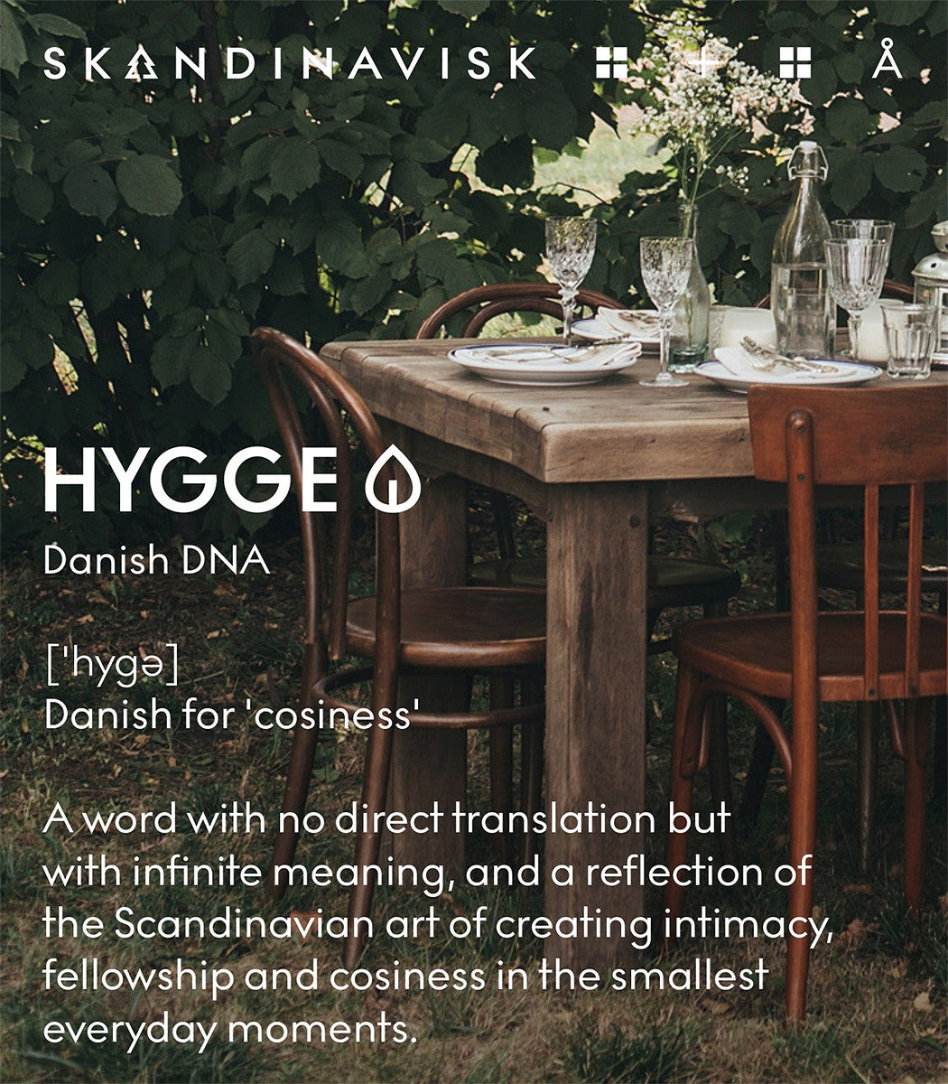 A rustic outdoor table with wooden chairs is set for dining amid lush greenery. The image features text describing hygge—the Danish art of coziness and everyday intimacy—accompanied by the Skandinavisk Giftset Scented Candles Hibernate, which includes three mini candles: Skog, Koto & Hygge, each crafted from Swedish rapeseed wax.