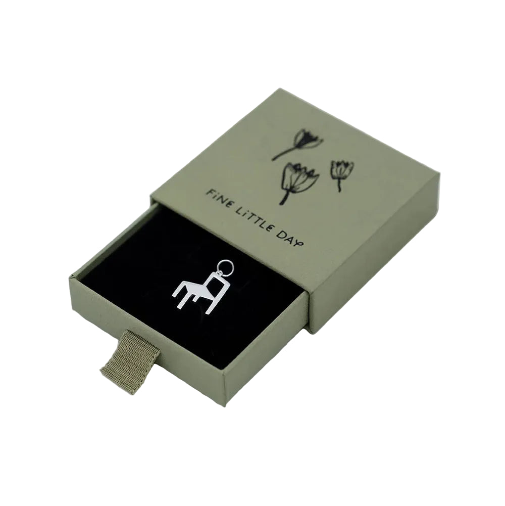 A small silver Chair Charm from Fine Little Day, shaped like a chair, is displayed in an open green box adorned with simple flower illustrations and labeled FINE LITTLE DAY, echoing the delicate design characteristic of recycled silver jewelry.