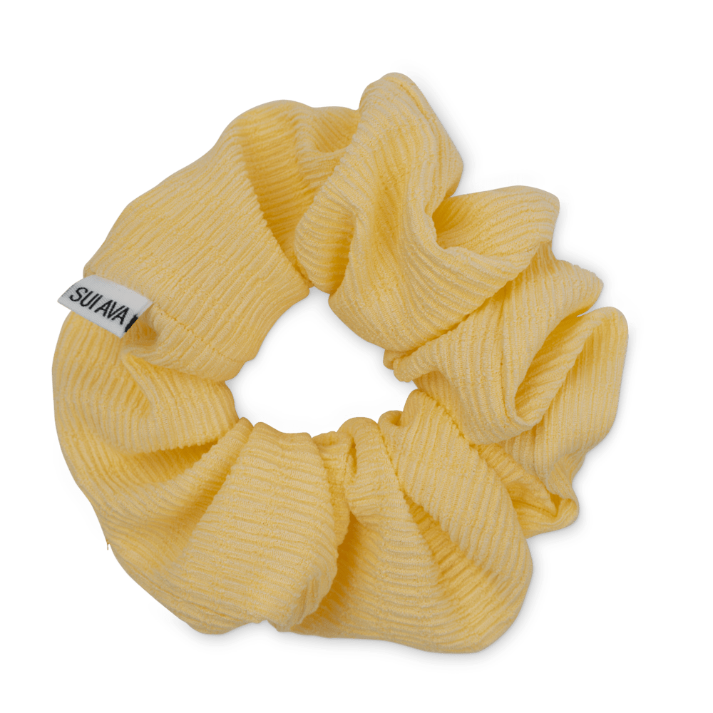 The Ea Zig-Zag Scrunchie in Blazing Yellow by Sui Ava is a ribbed fabric hair accessory with a small white SUIAVA tag, bringing a vibrant touch to any look when styled on a light gray background.