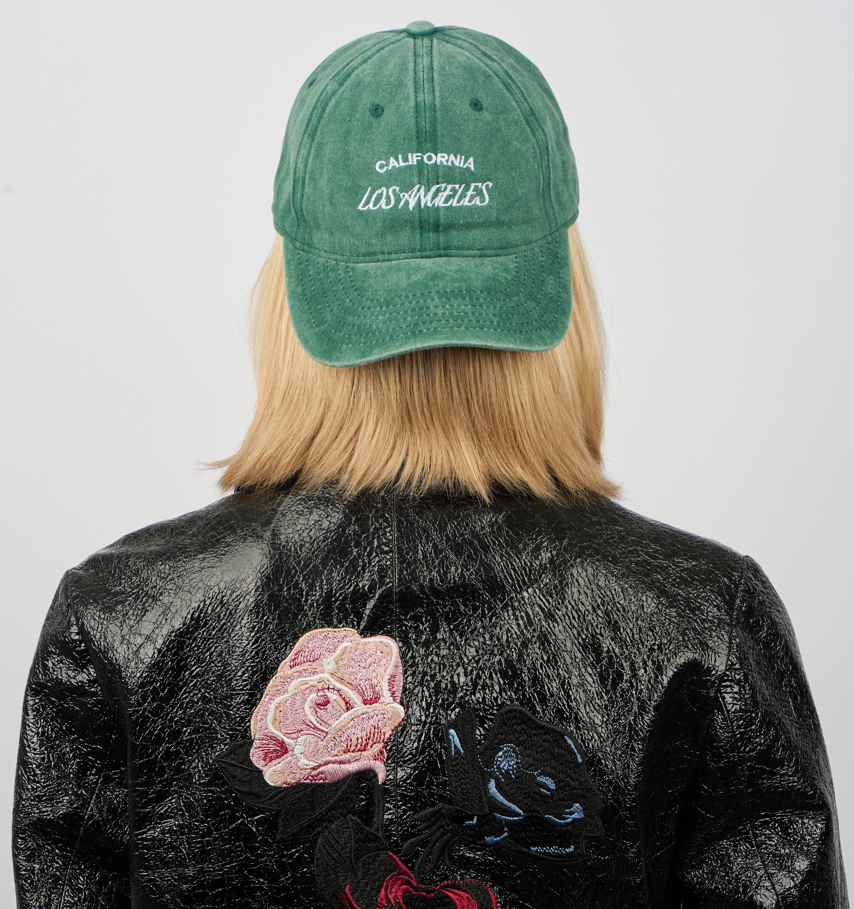 A person with shoulder-length blonde hair, seen from behind, wears the Sui Ava Everyday Cap, Los Angeles in Washed Green with an adjustable strap, paired with a black leather jacket featuring embroidered pink and blue flowers on the back.