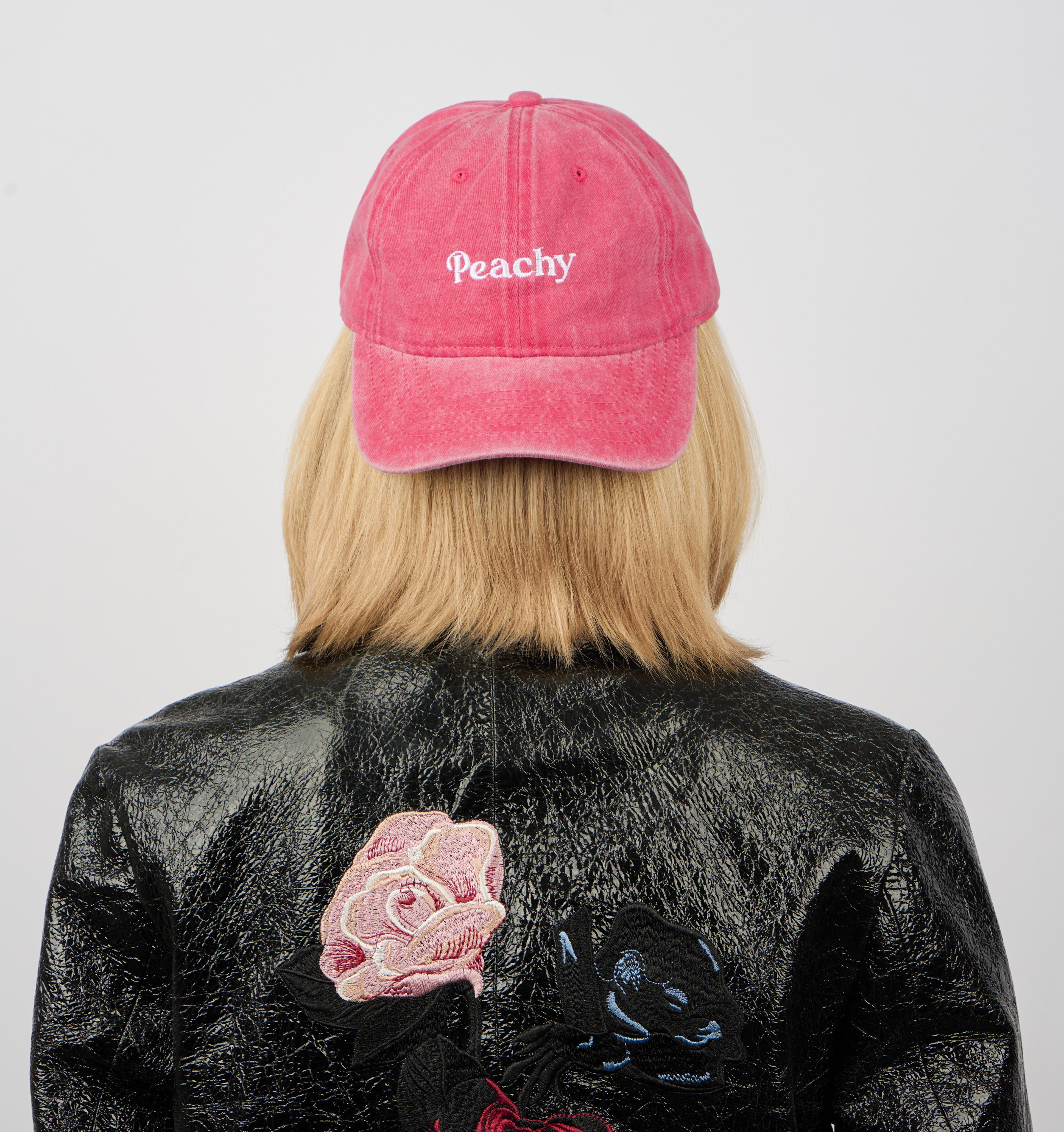 A person with blonde hair, seen from behind, wears Sui Ava’s Everyday Cap in Peachy and a black textured jacket with rose embroidery, against a plain white background.