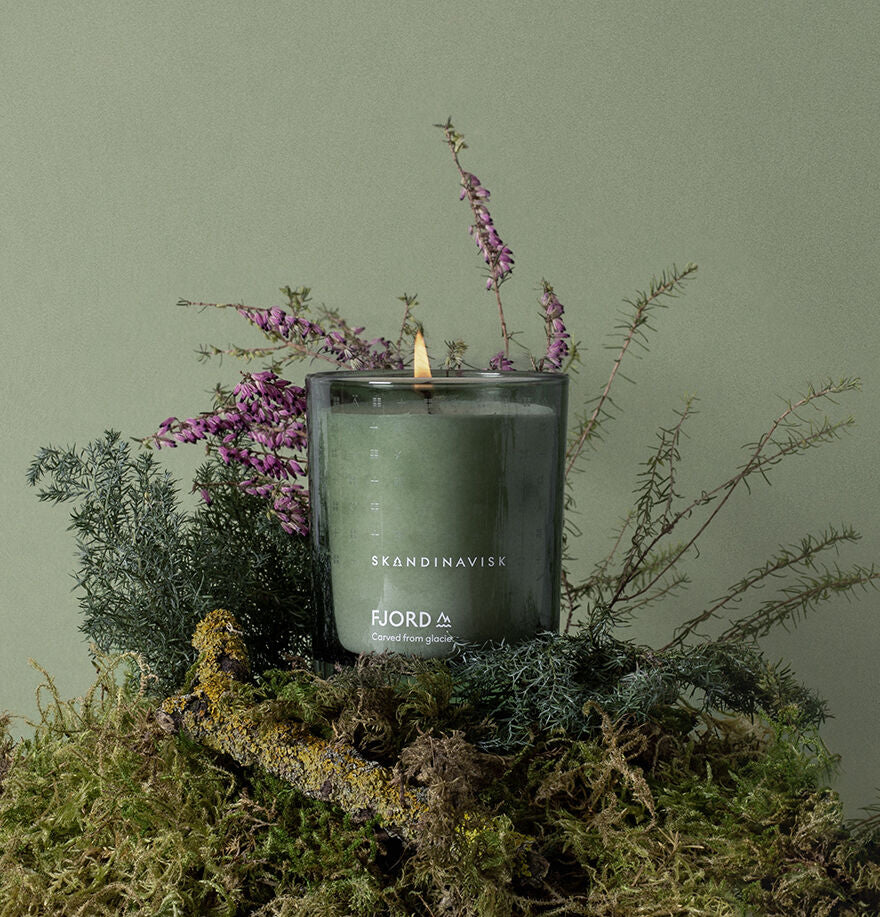 A Skandinavisk Scented Candle Fjord 50h, 200g made from Swedish rapeseed wax is lit on moss, surrounded by greenery and purple flowers, set against a green background.