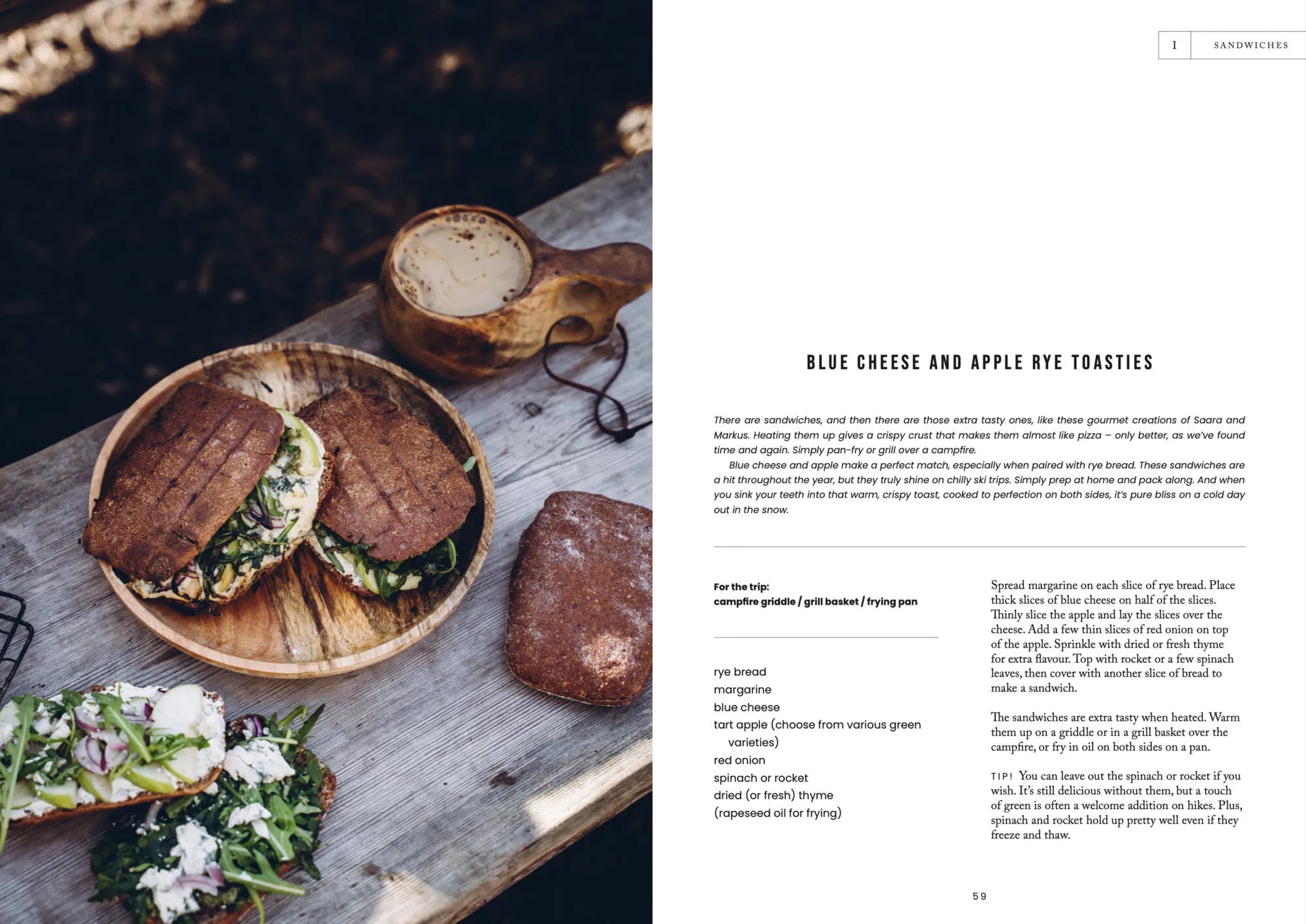 A wooden platter displays two rye bread toasties stuffed with blue cheese, apple, spinach, and onions next to a steaming mug of coffee on a rustic picnic table. Nearby, a plate holds an open-faced toastie—an inviting spread that sparks inspiration for vegetarian campfire meals deep in the wilderness. The right page features a recipe from “Food Over the Campfire – Recipes for Day Hikes and Wilderness Treks” by Cozy Publishing.
