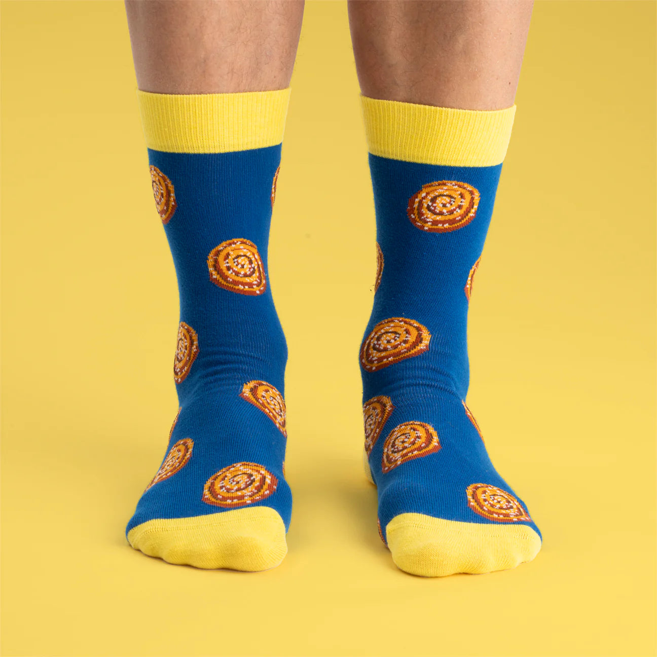A person wearing Svensk Husman Socks, Cinnamon Bun—blue seamless socks with cinnamon roll patterns plus yellow cuffs, heels, and toes—stands on a yellow background, capturing the cozy spirit of Swedish fika.