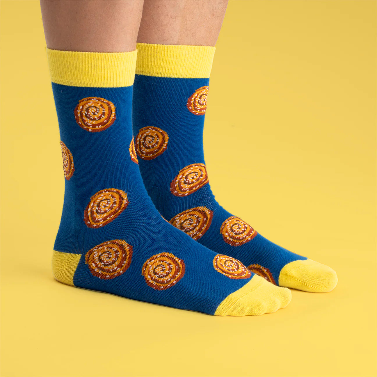 A person wears Svensk Husman Socks' Cinnamon Bun socks—featuring playful cinnamon roll patterns and blue-yellow accents—while standing on a vibrant yellow background.
