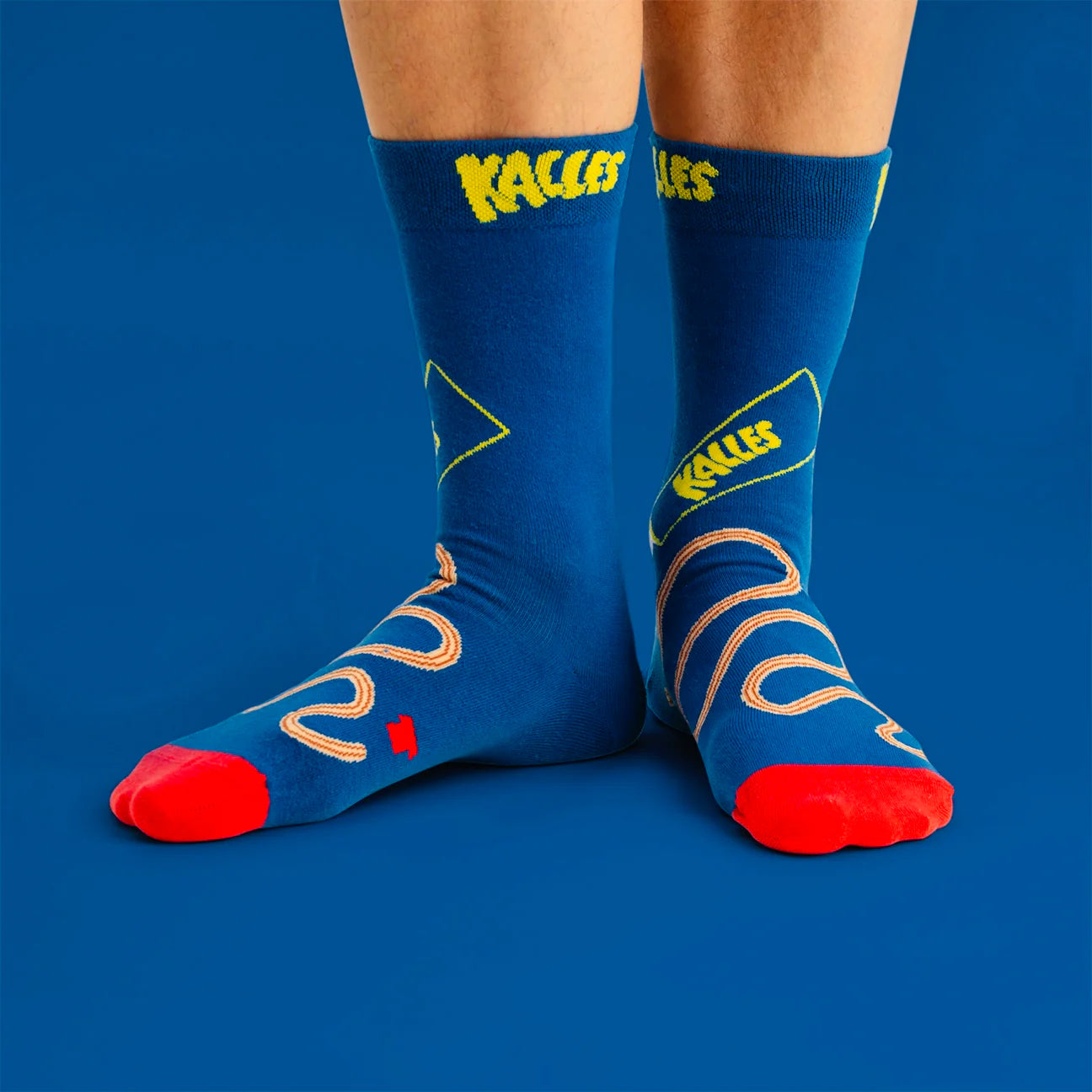 Feet in blue Svensk Husman Socks, Kalles Caviar The Original Special Edition—featuring red toes, yellow designs, “Kalle's Caviar,” abstract squiggles, and “Kalles”—shown on a solid blue background.