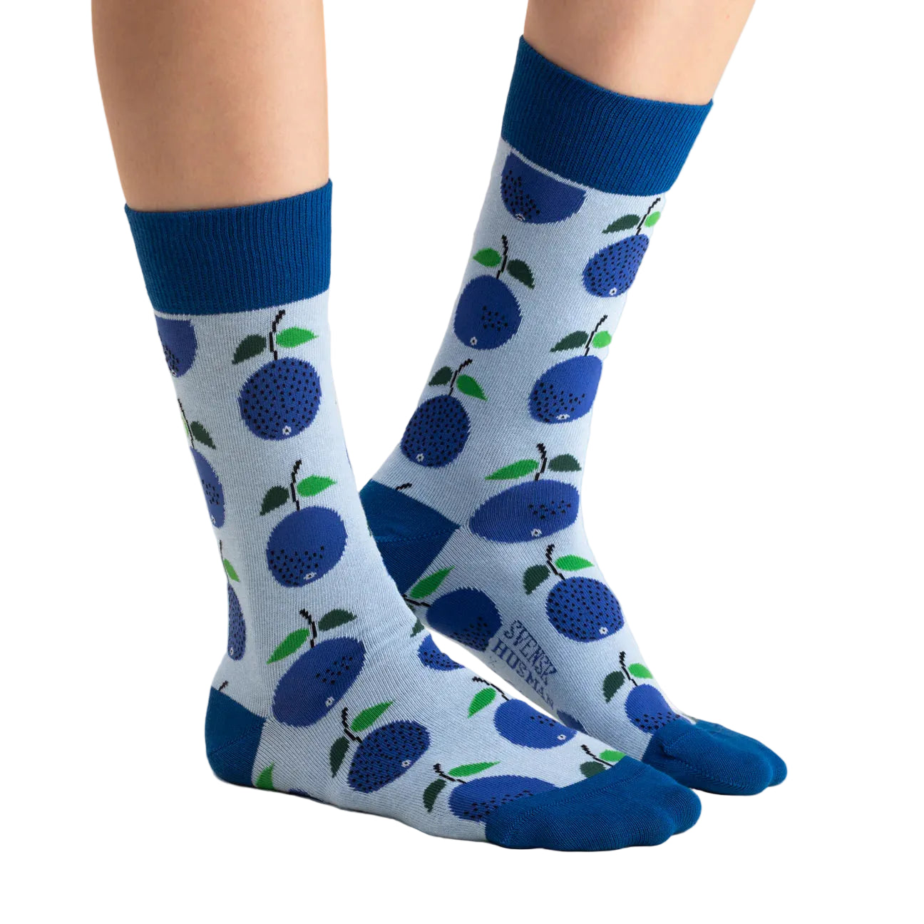 A person wearing Svensk Husman Socks' Stig L Prunus: light blue socks with dark blue cuffs, toes, and heels, featuring a Swedish-inspired design of large blue plums with green leaves and black dots.