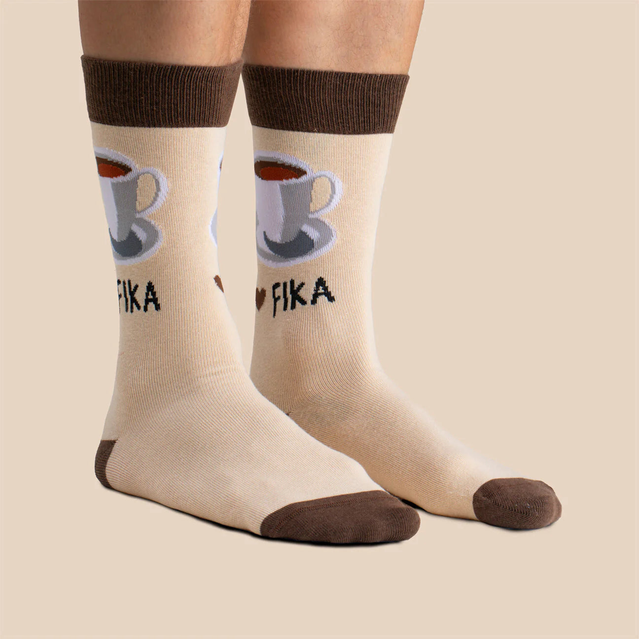 A person wears beige Svensk Husman Socks, Coffee Cup Fika—featuring brown heels, toes, and cuffs, a coffee cup graphic, and "I ♥ FIKA," inspired by Swedish culture—while standing on a neutral background.