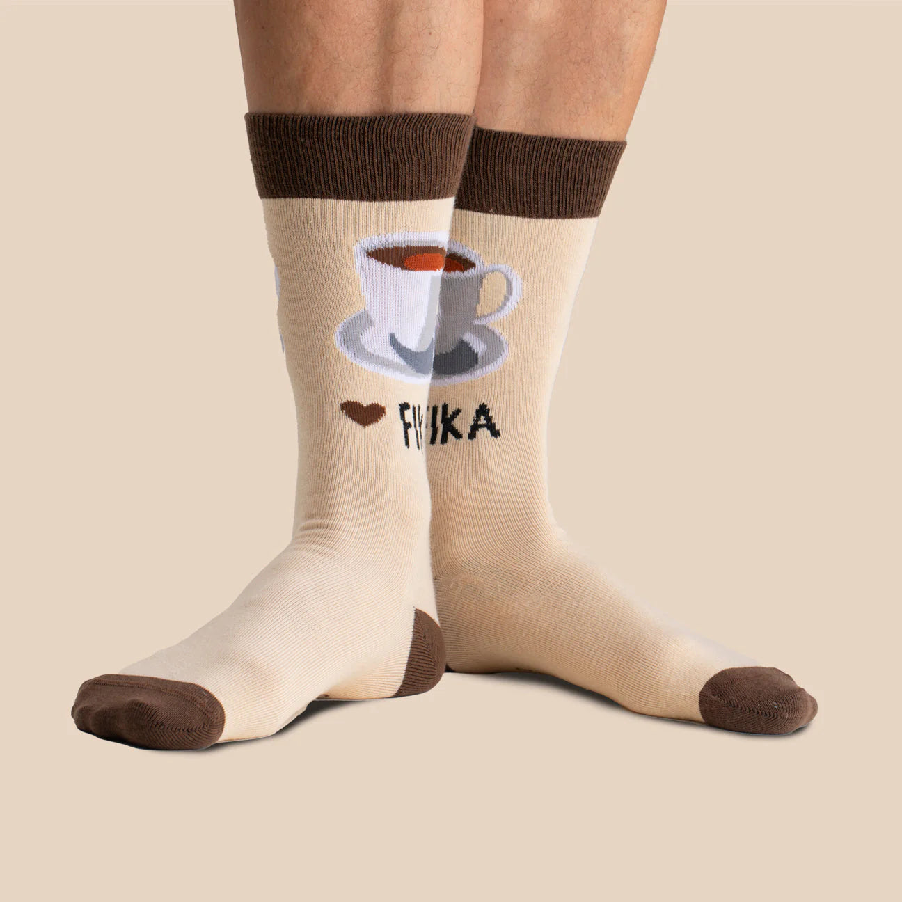 Svensk Husman Socks’ Coffee Cup Fika features beige socks with brown accents, a coffee cup and heart pattern, plus "FIKA" near the ankle—a cozy Swedish-inspired design.